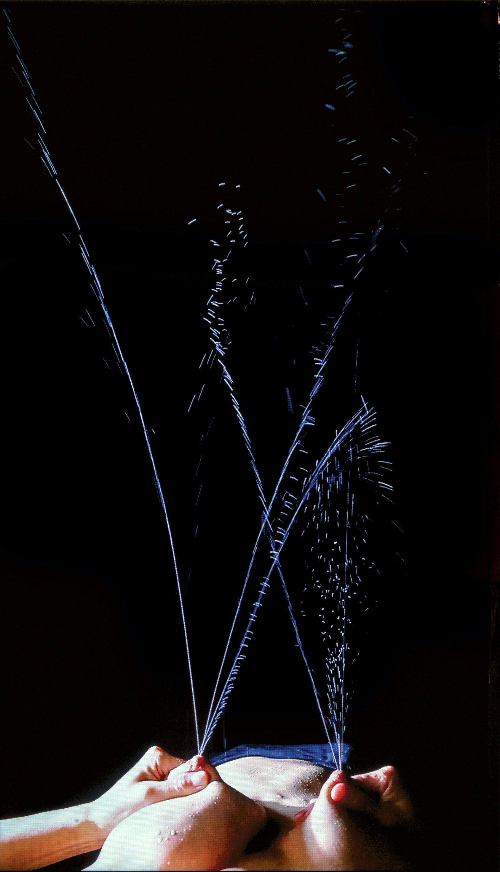 The subject's hands squirt liquid from their breasts emulating the action of a water fountain. The body is lays on the floor surrounded by a black backdrop.