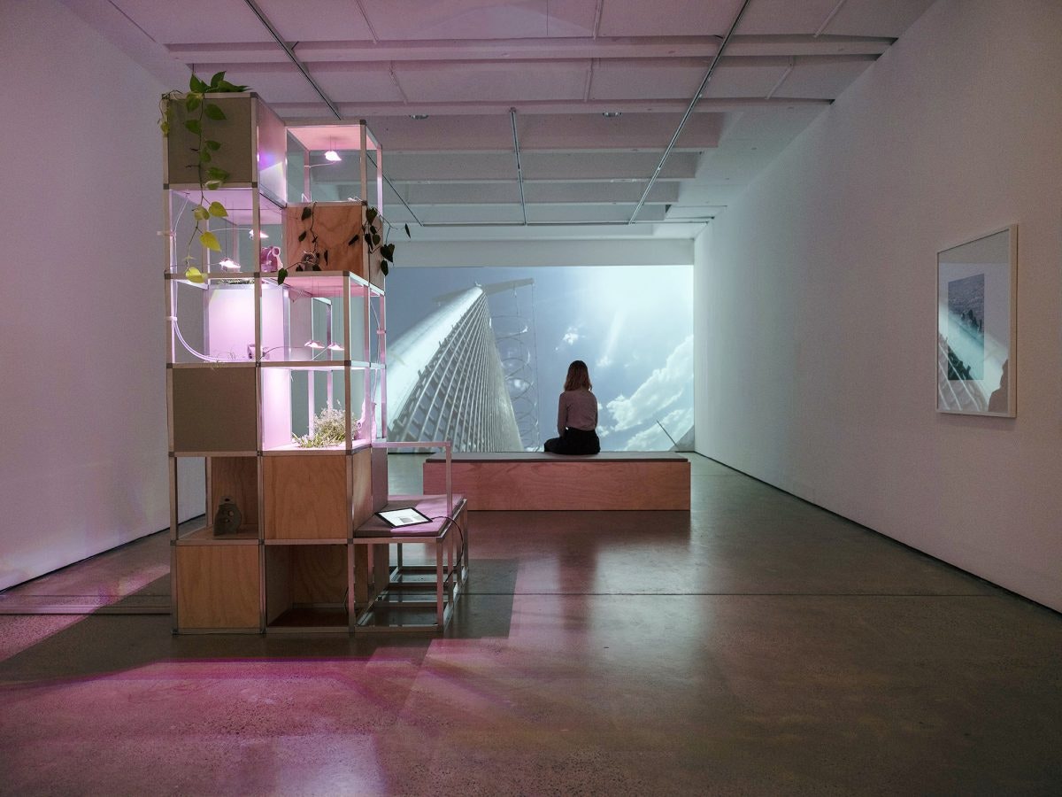 A female-presenting figure sits on a wooden bench, facing a projection of a tower and a blue sky with clouds.