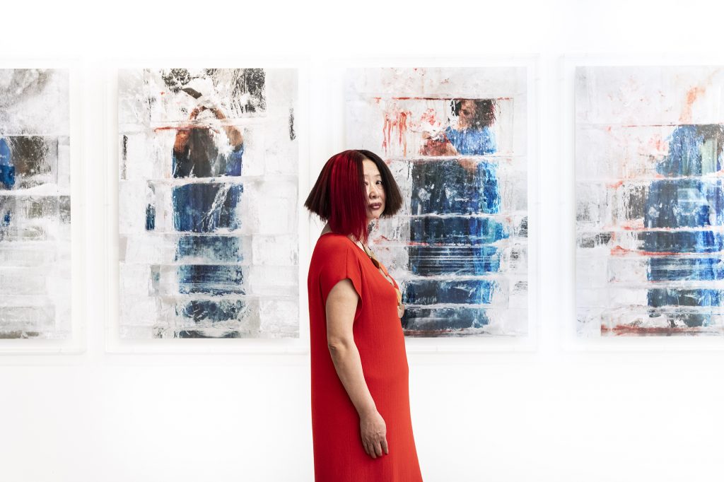 A female-presenting Han Chinese figure with dark-red dyed hair and a bright red shift dress stands in front of four printed canvases in an art gallery, looking towards the camera with a relaxed expression.