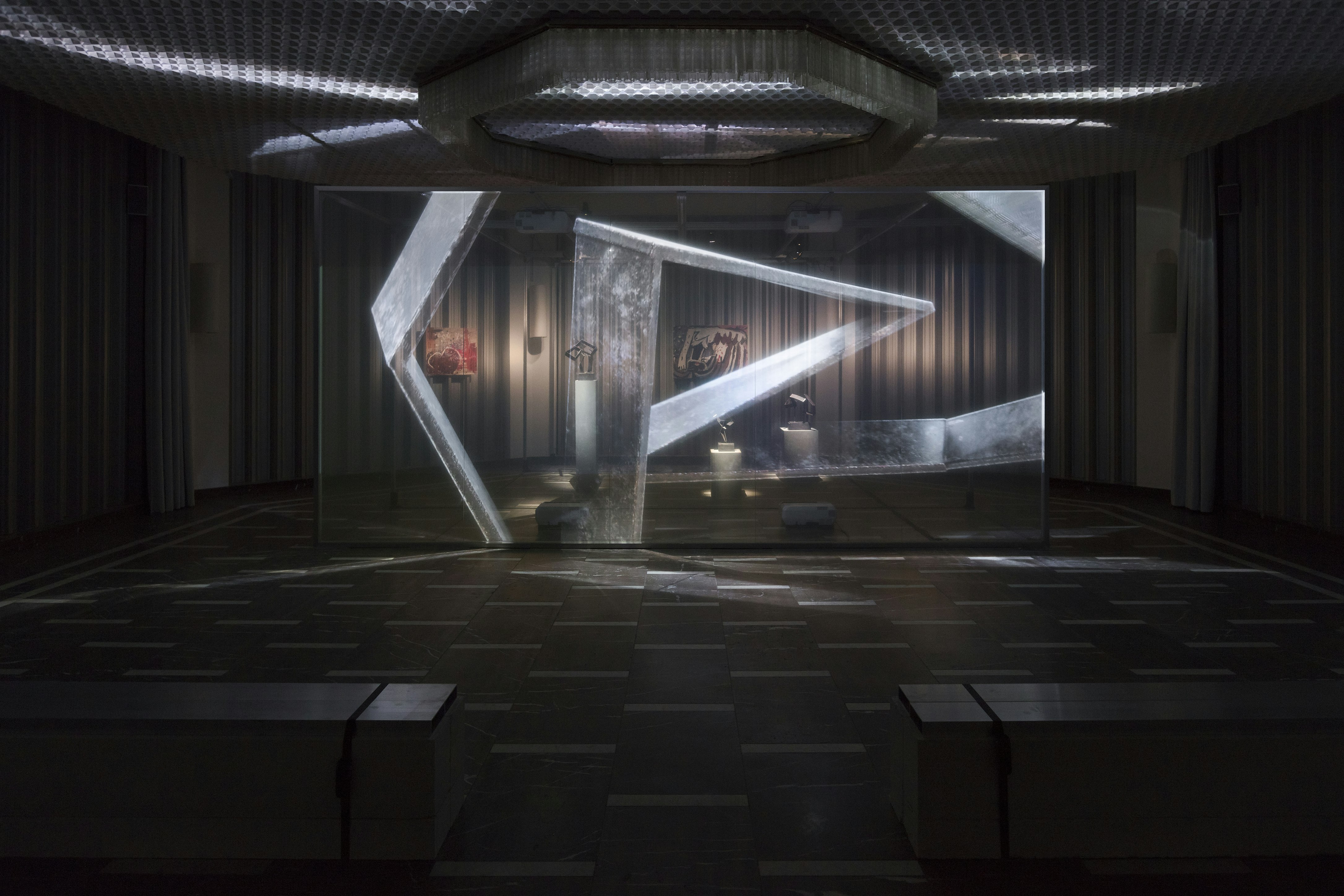 A darkly lit, futuristic gallery space with a film projected onto a large transparent screen.
