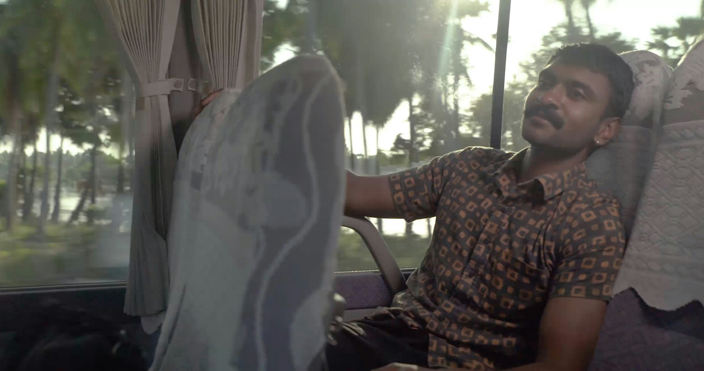 A brown South Asian male-presenting figure sits on a bus in a printed button up shirt, while a sunny tropical scene is shown through the bus window.