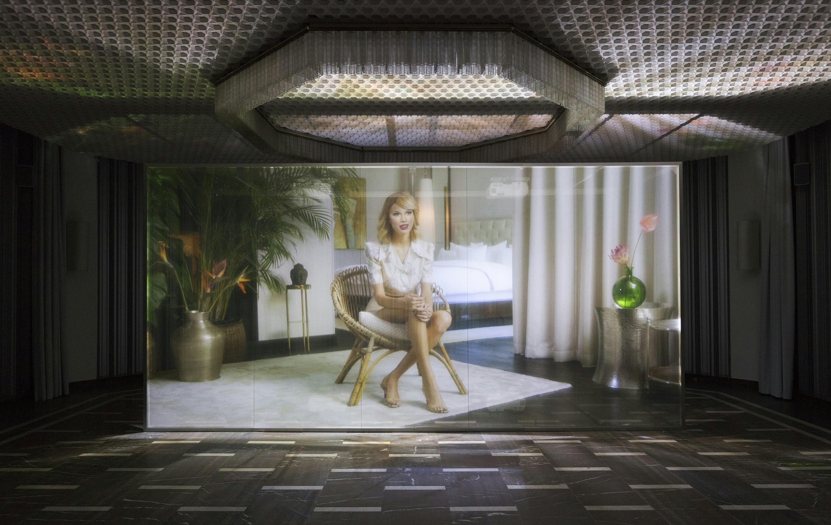 A digitally generated projection of a blonde Caucasian woman closely resembling popstar Taylor Swift, sitting cross legged in a rattan chair, surrounded by potted plants. This image is projected onto a large transparent screen in a darkly lit exhibition space.