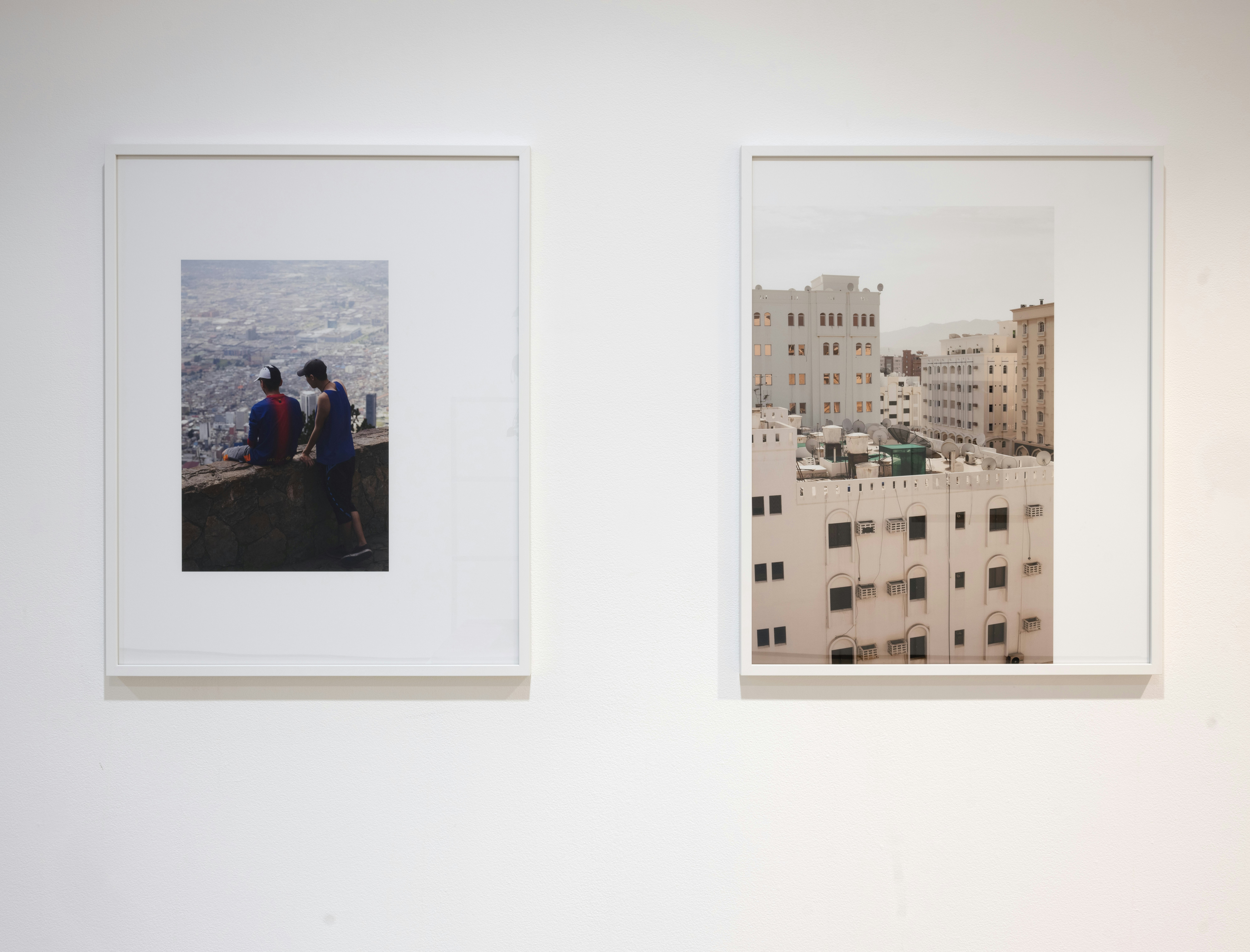 Two photographic prints in white frames on a gallery wall. The left print is of two male-presenting figures looking over a city, the right is a shot of tall white residential buildings.