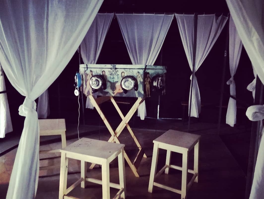 A dark room surrounded by white knotted curtains, with a bioscope-inspired sculpture in the centre, surrounded by three wooden stools.