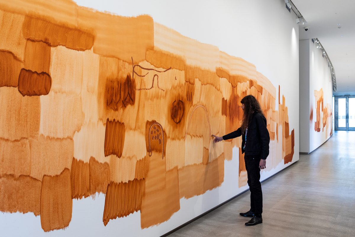 A male-presenting figure with long curly hair touches a wall painting which is comprised of textured brown brushstrokes.