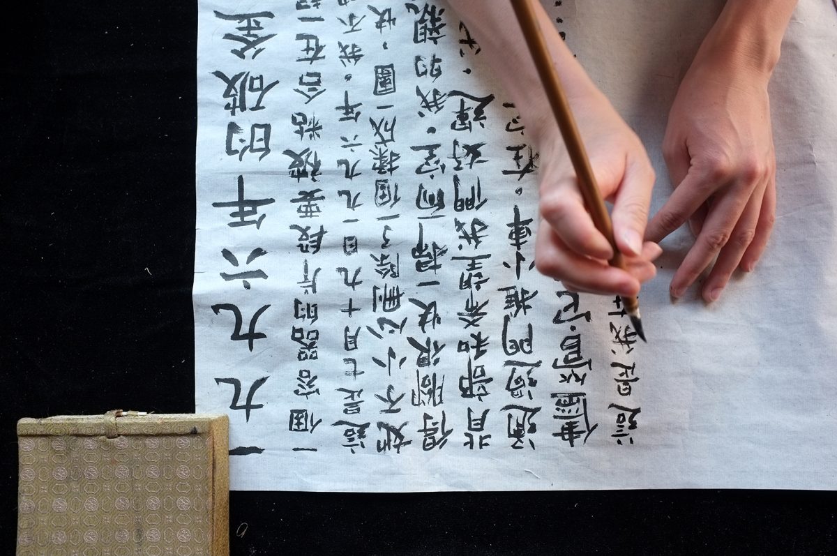 Flatlay view of two hands holding a calligraphy brush, drawing Chinese characters on a white sheet of paper.