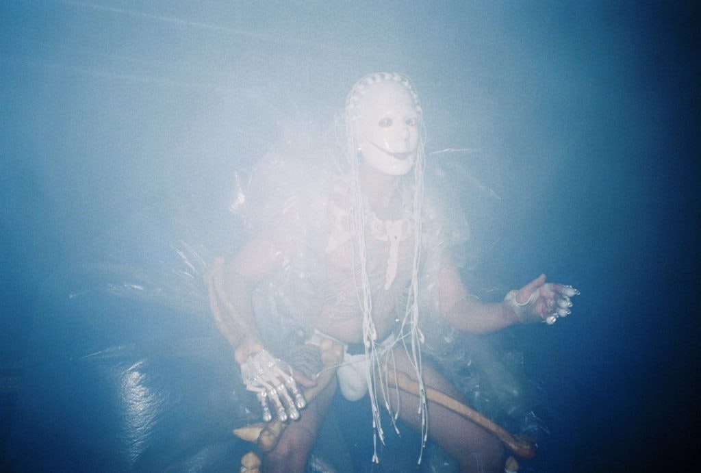 A figure wearing a white mask with braided decorations, dancing in the dark.