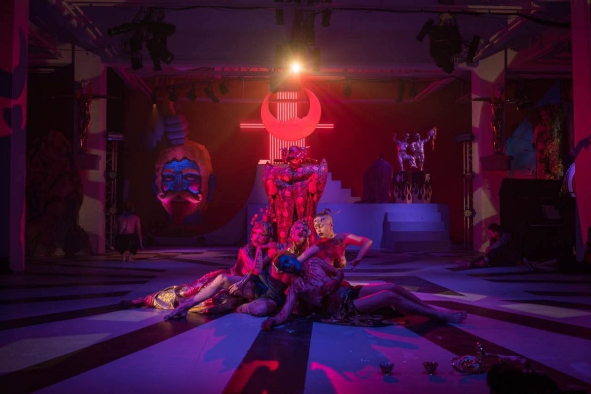 Four dancers sitting at the base of a sculpture in a red/purple-lit room, with a spotlight shining on them.
