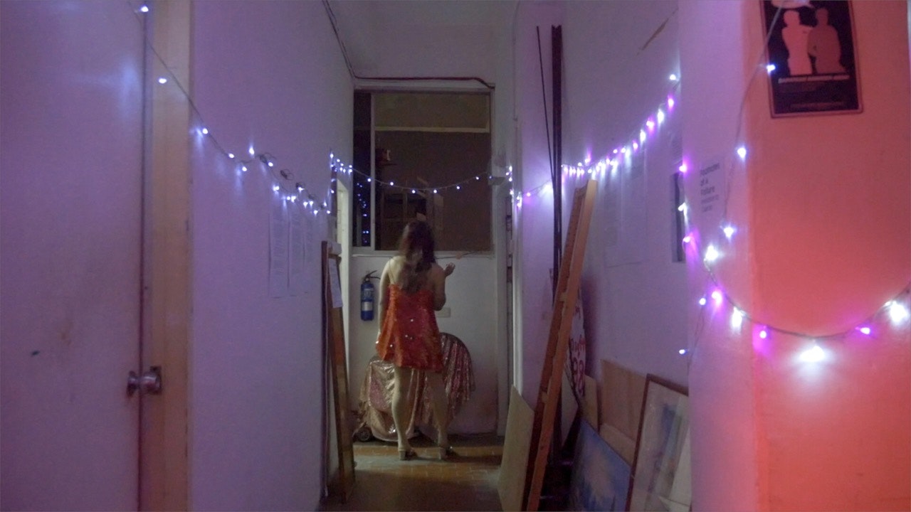 A female-presenting figure in a red dress stands at the end of a hallway with her back to us, looking out a window. The hallway is strung with fairy lights.