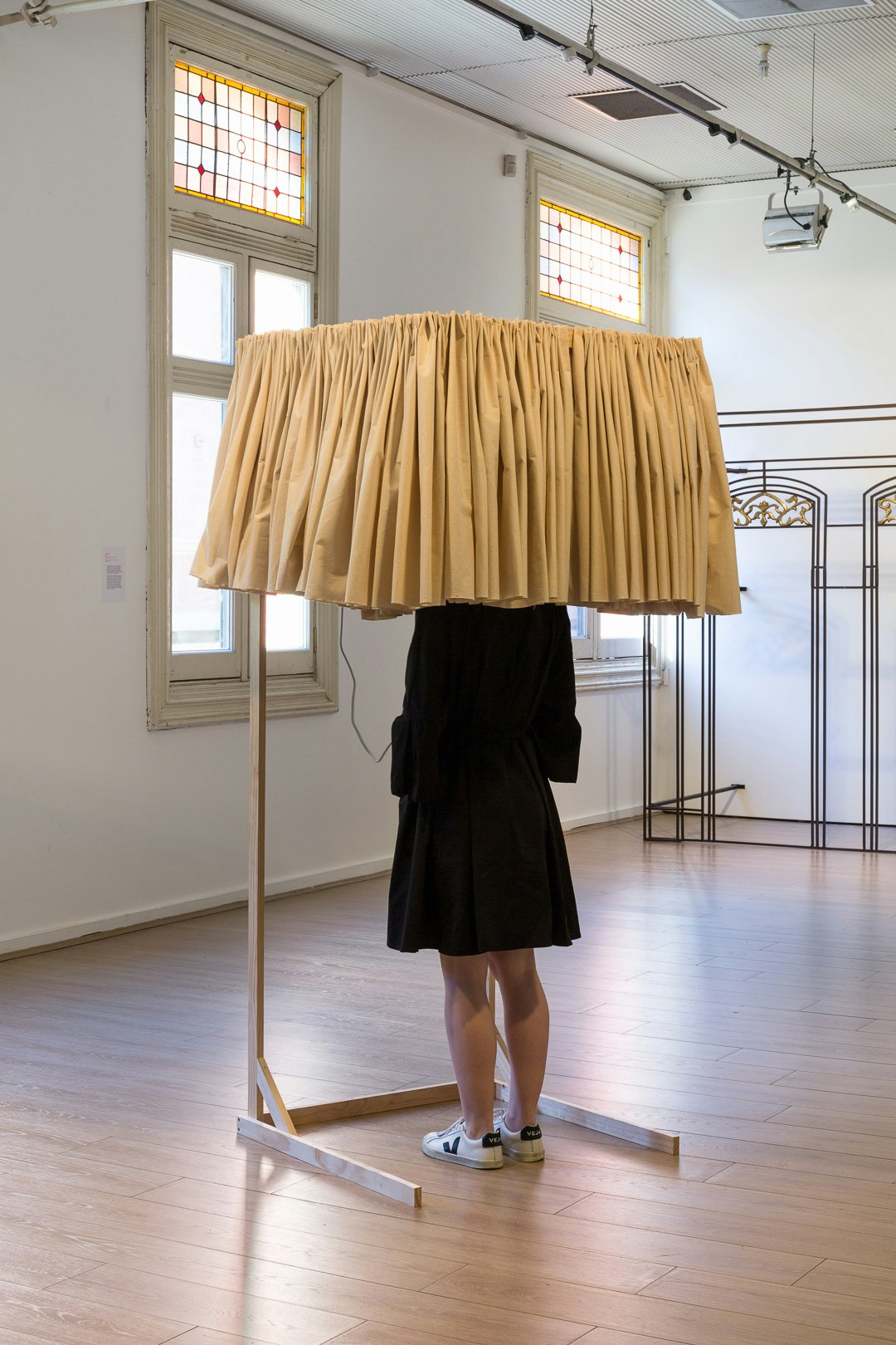 A figure in a black dress and white sneakers stands with their head shrouded by a standing curtain installation.