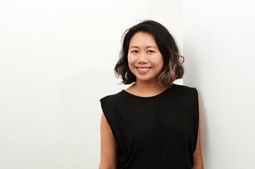 A woman of East Asian descent with short ombre black hair stands against a white wall, wearing a black shift dress.