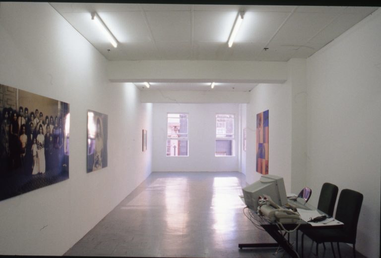 A view of a white gallery space with some photographs and paintings hung on the walls, lit by fluorescent lights and natural light coming in from two nearby windows.