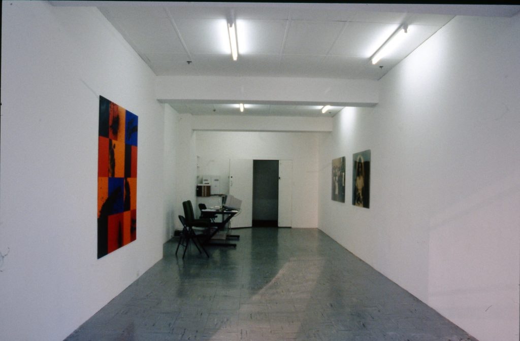 A white gallery space with artworks on the walls. The closest artwork is a series of technicolour squares in red, orange and blue.