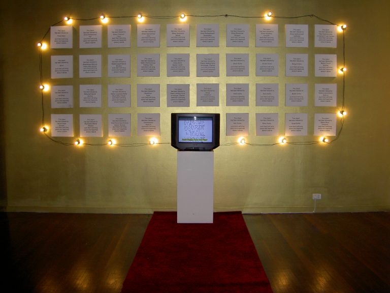 A television on a plinth, surrounded by a wall of 38 plaques with written text, framed by yellow fairy lights.