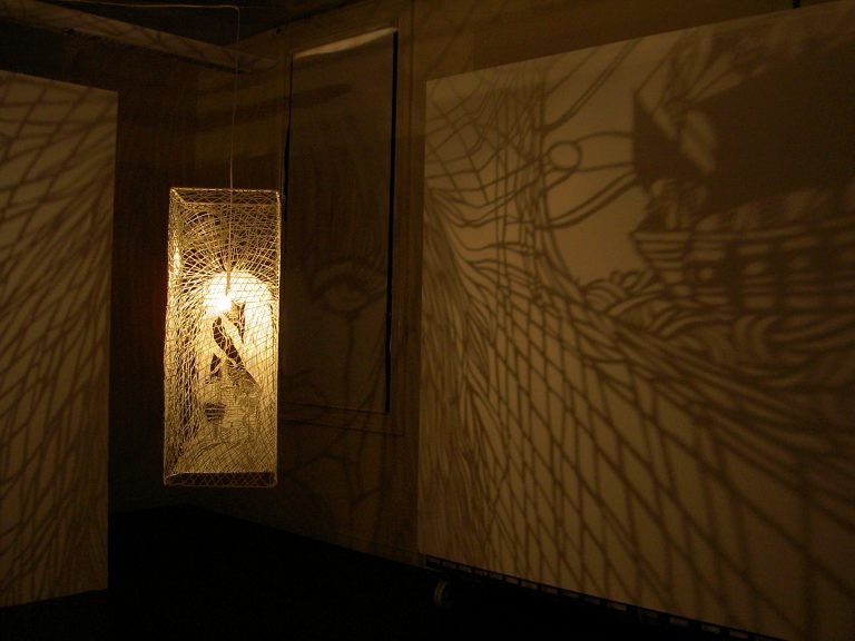 A yellow light behind a glass panel in a dark room, throwing patterned shadows onto the right wall.