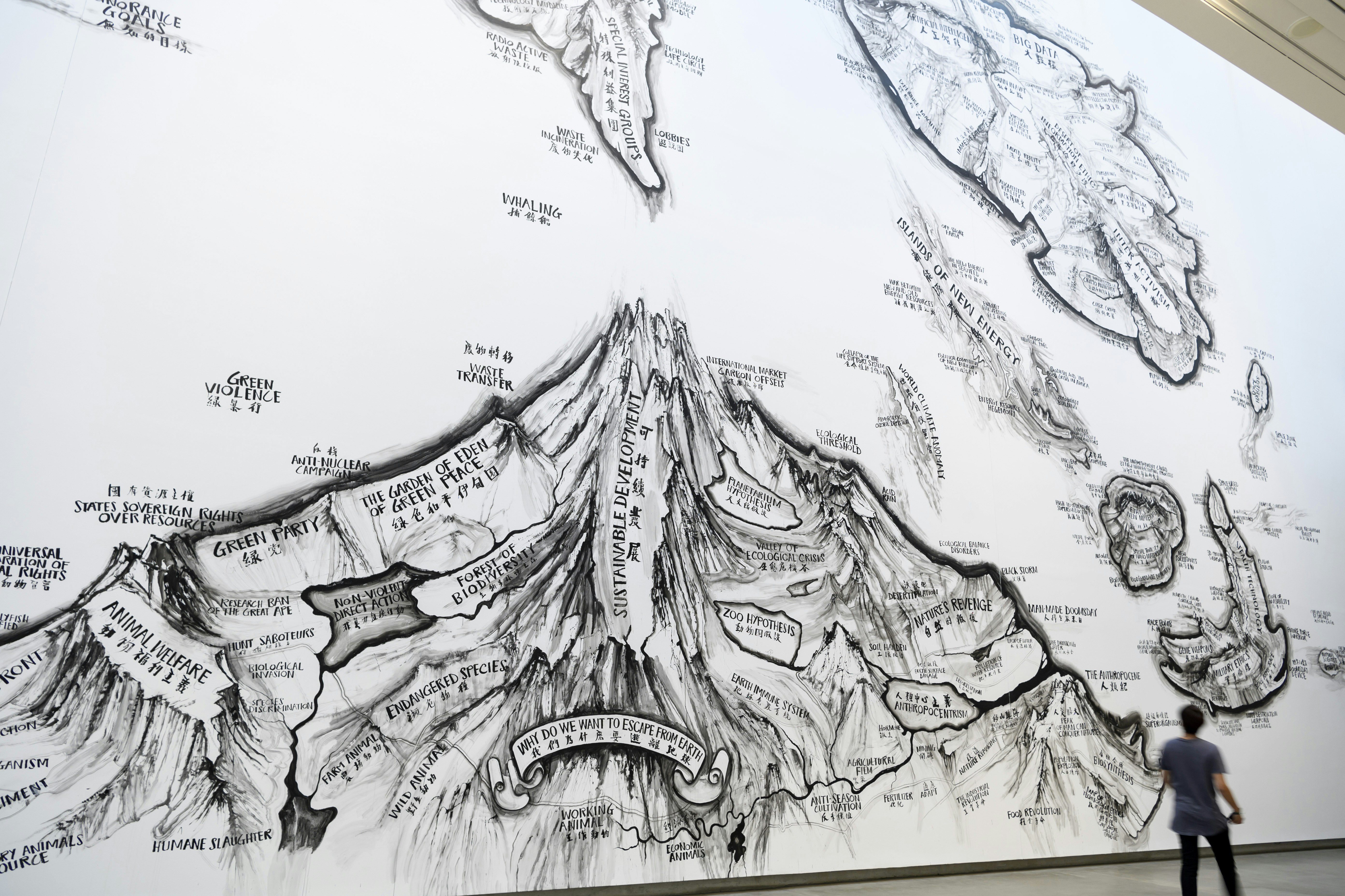 A figure walks past a grand wall installation of a topographical drawing resembling a mountain and some surrounding islands.
