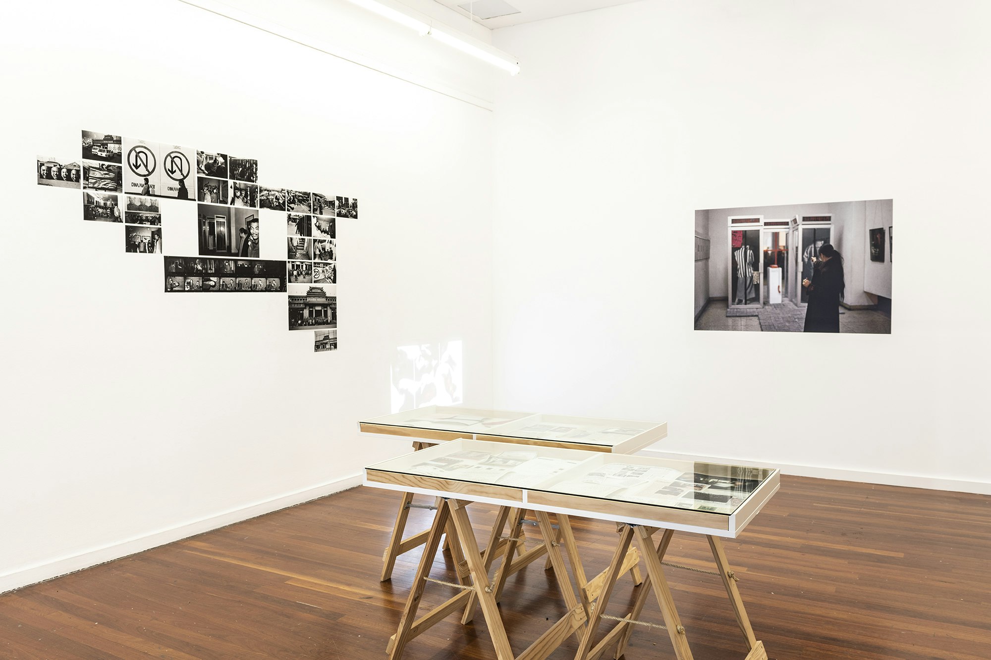 A white gallery space with two glass display cabinets, a projector stacked on concrete blocks facing a wall and a selection of black inkjet photographs pasted up on the wall
