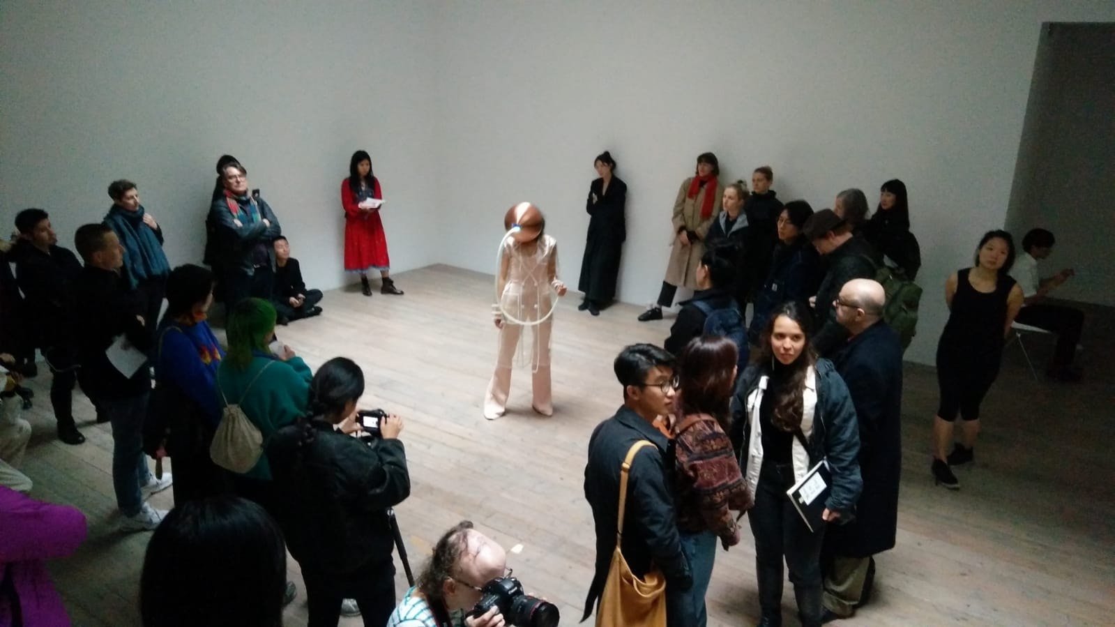 A performer wearing a futuristic bronze helmet and sheer jumpsuit, surrounded by a circle of patrons in a gallery.
