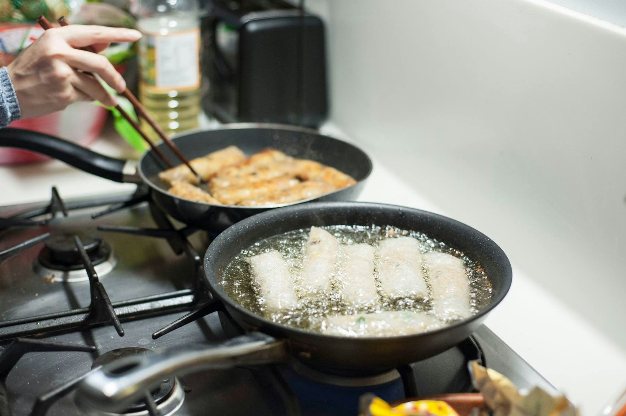 Two frying pans filled with oil and chả giò (Vietnamese spring rolls). A hand holding chopsticks turns one of the spring rolls over in the pan.