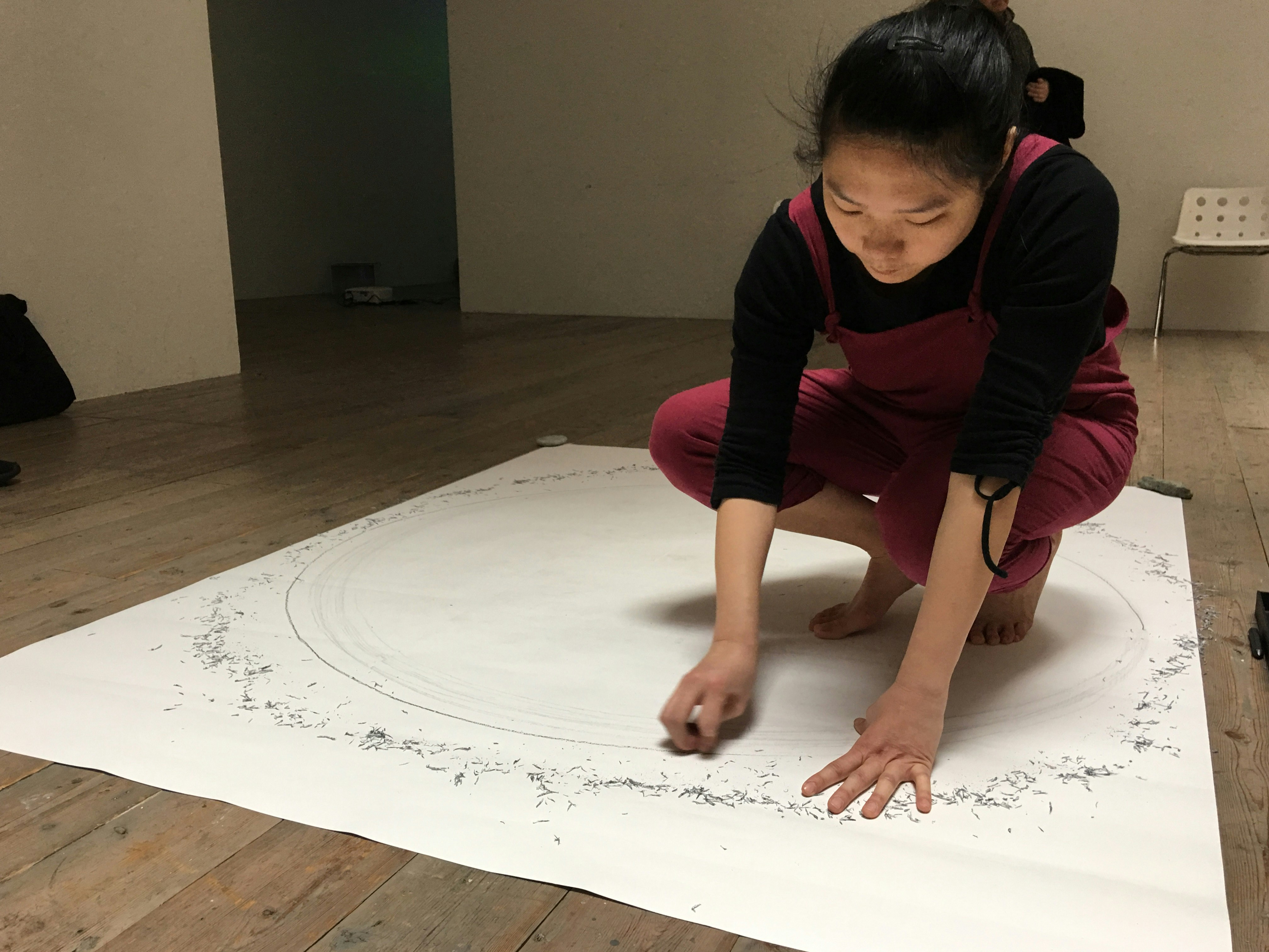 A female-presenting artist of East Asian descent squats on a large piece of paper, drawing a circle around herself.