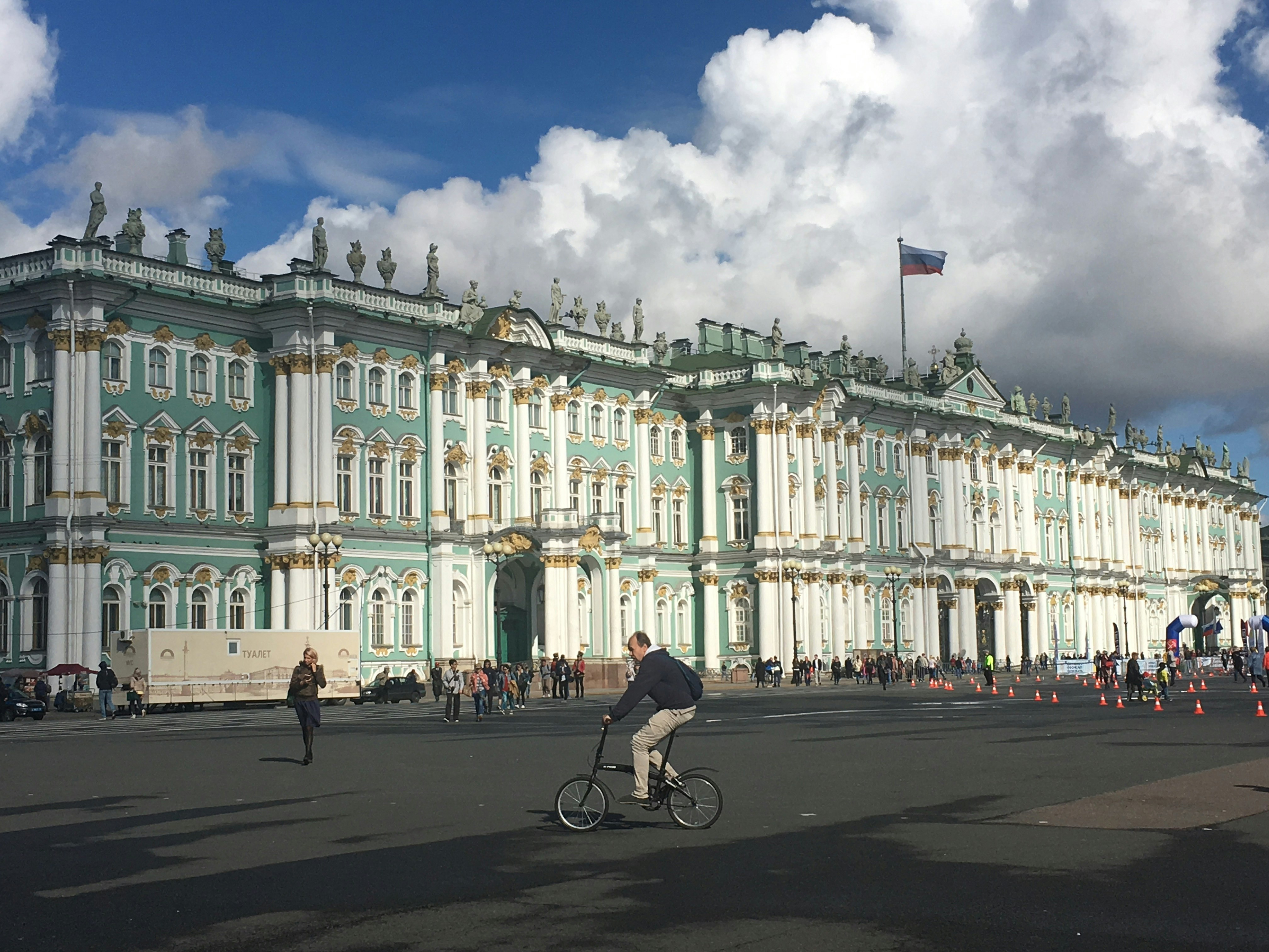 A male-presenting figure rides a bicycle outside the front of State Hermitage Museum, a regal teal green building with gold architectural embellisments.
