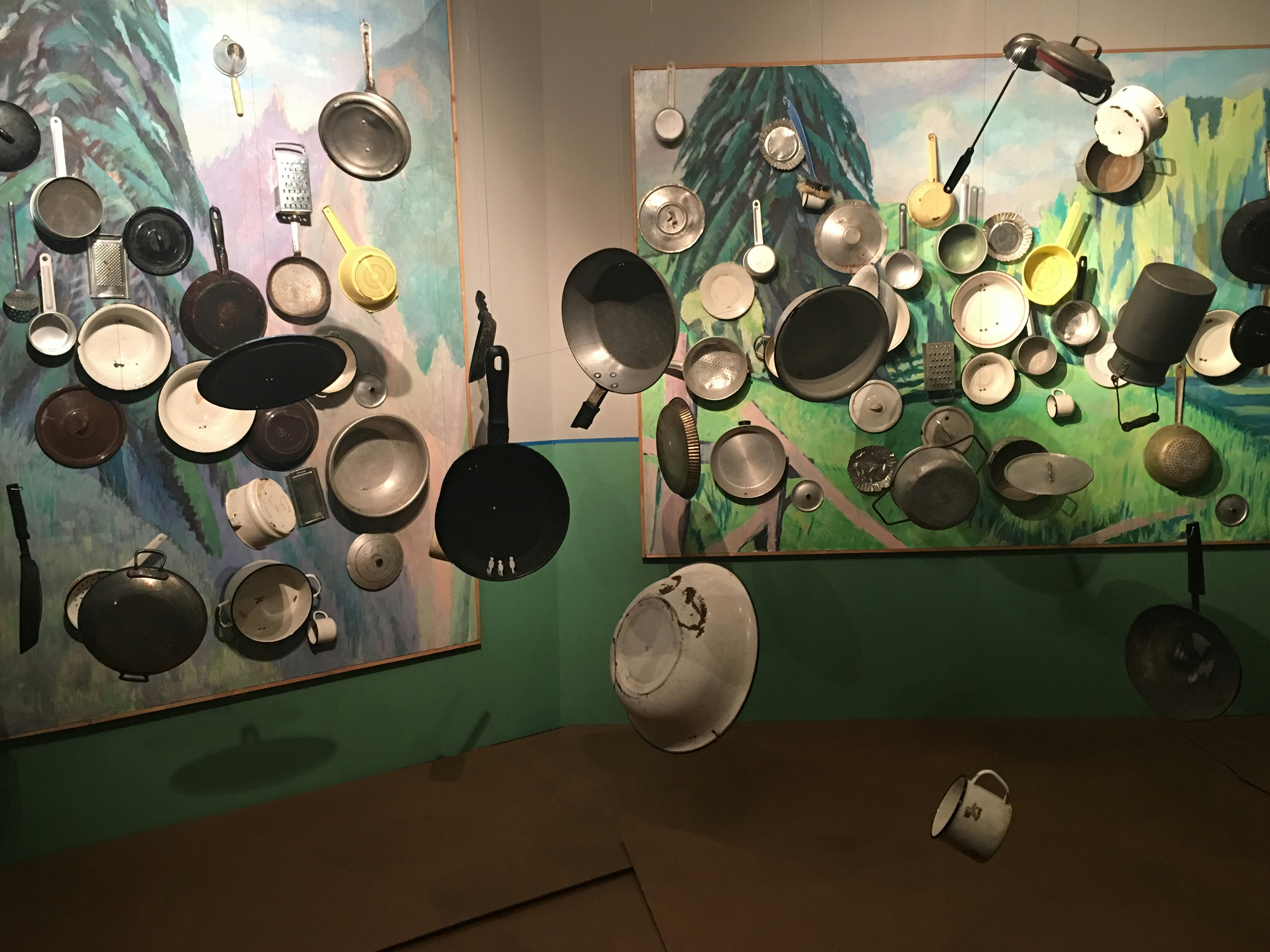 A series of suspended pots, pans and other enamel kitchenware, hanging in front of two large impressionistic landscape paintings.