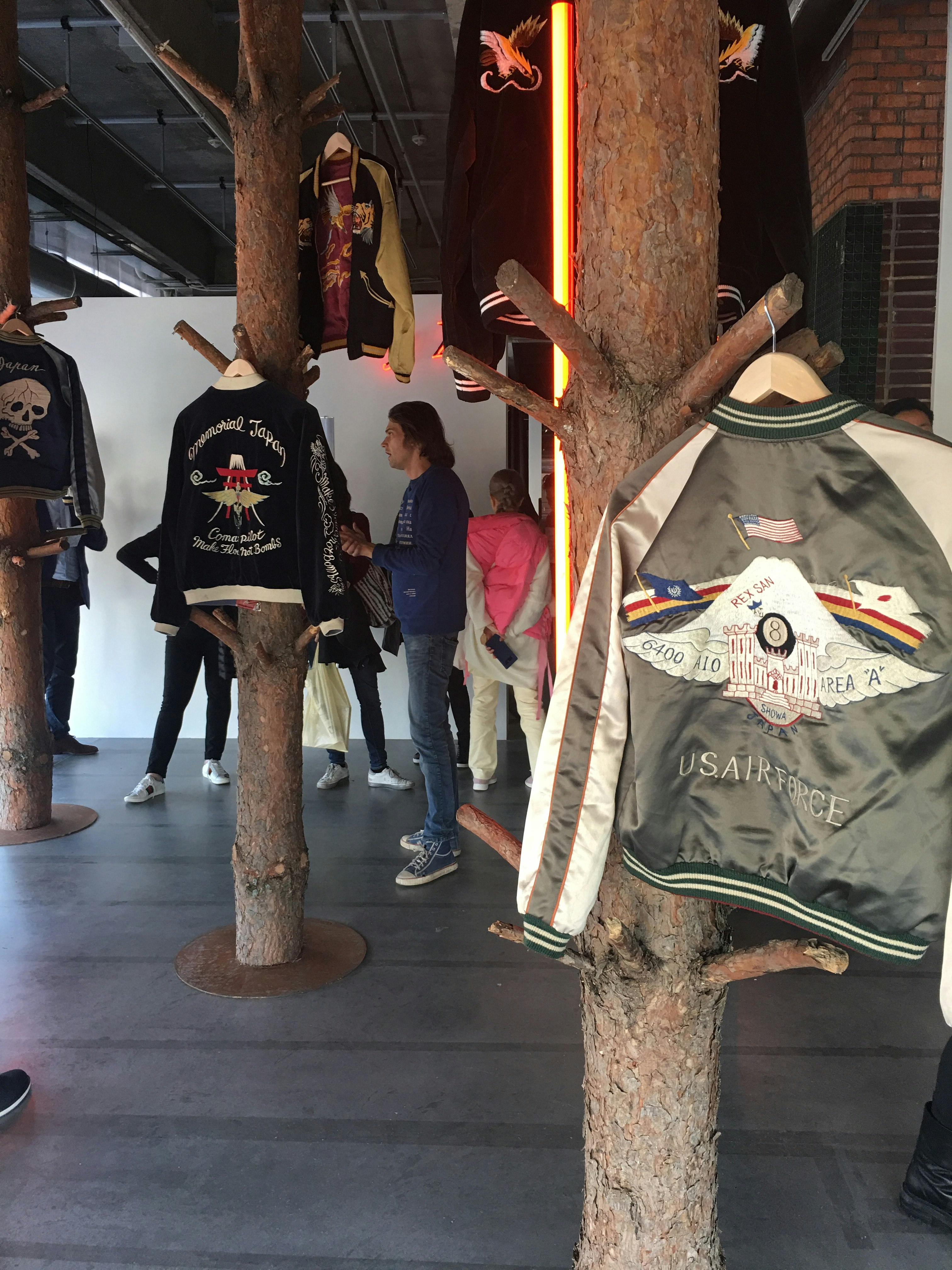 A U.S Air Force bomber jacket hanging on a tree branch in a gallery, surrounded by other embroidered bomber jackets hooked onto installed tree trunks.