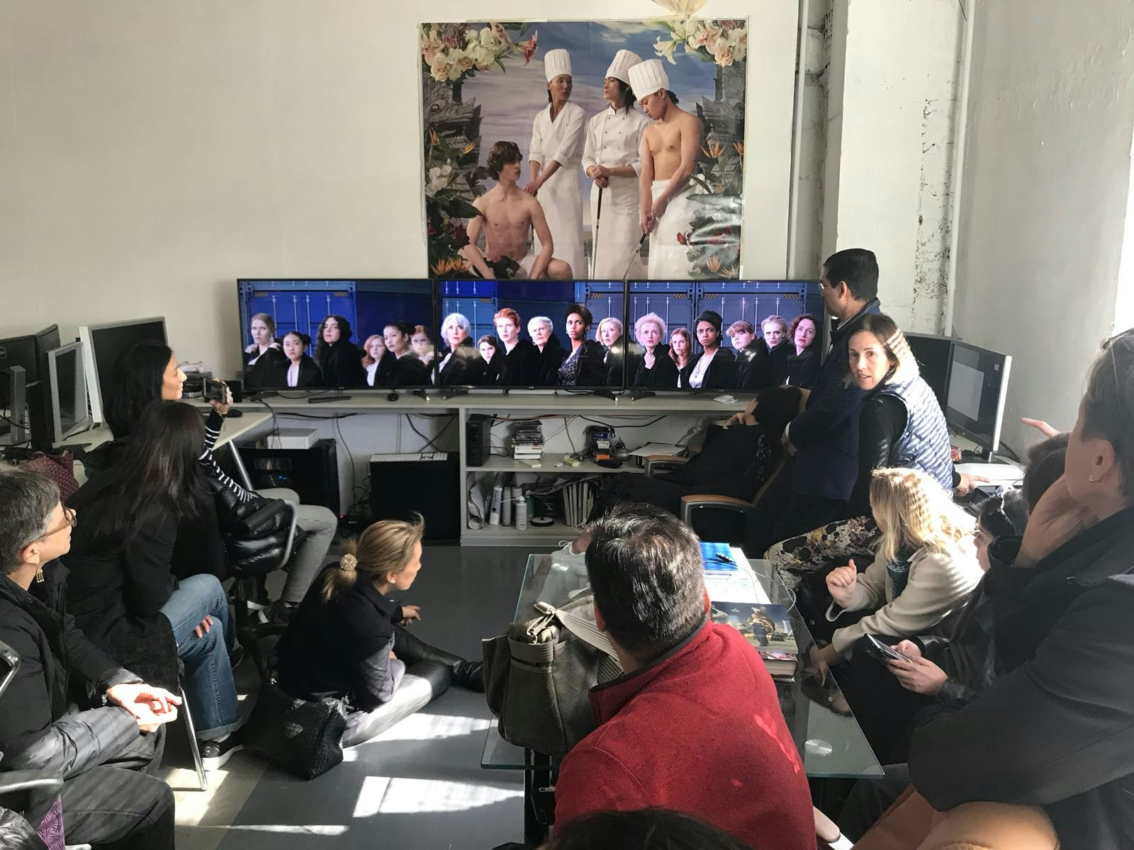 An audience observing three computer monitors lined side-by-side in an installation showing a line of people from the bust up. Behind the monitors is a large painting of three chefs and a shirtless seated figure, surrounded by white roses and lilies.