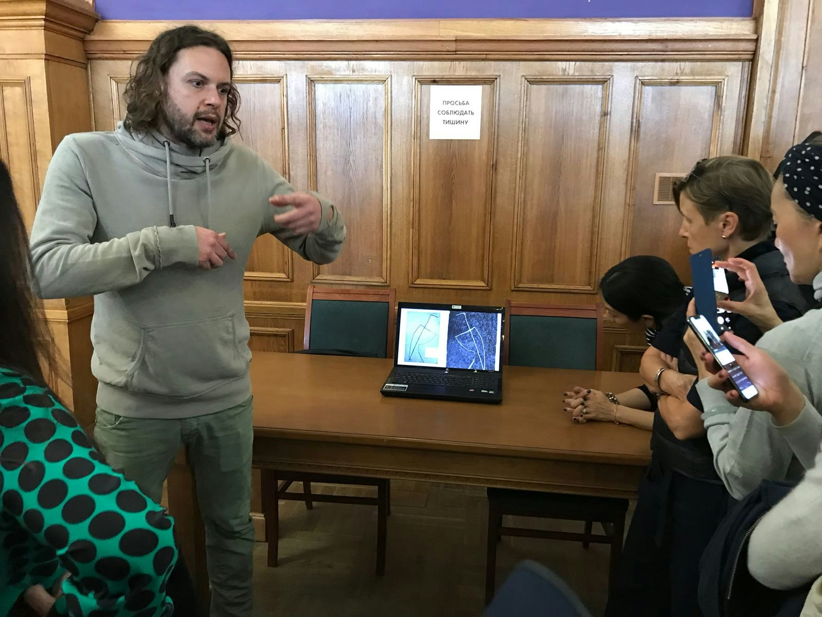 A male-presenting figure in a grey hoodie and green chinos, with curly shoulder-length brunette hair and a beard, presenting a talk in front of an open laptop.