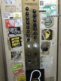 A panel of elevator call buttons surrounded by old peeling stickers inside an elevator car.