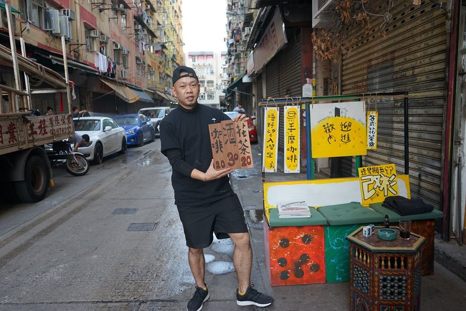 An East Asian male-presenting figure, wearing an all-black outfit and a backwards-facing black cap, holds up a handpainted sign on a back alley in Hong Kong. 