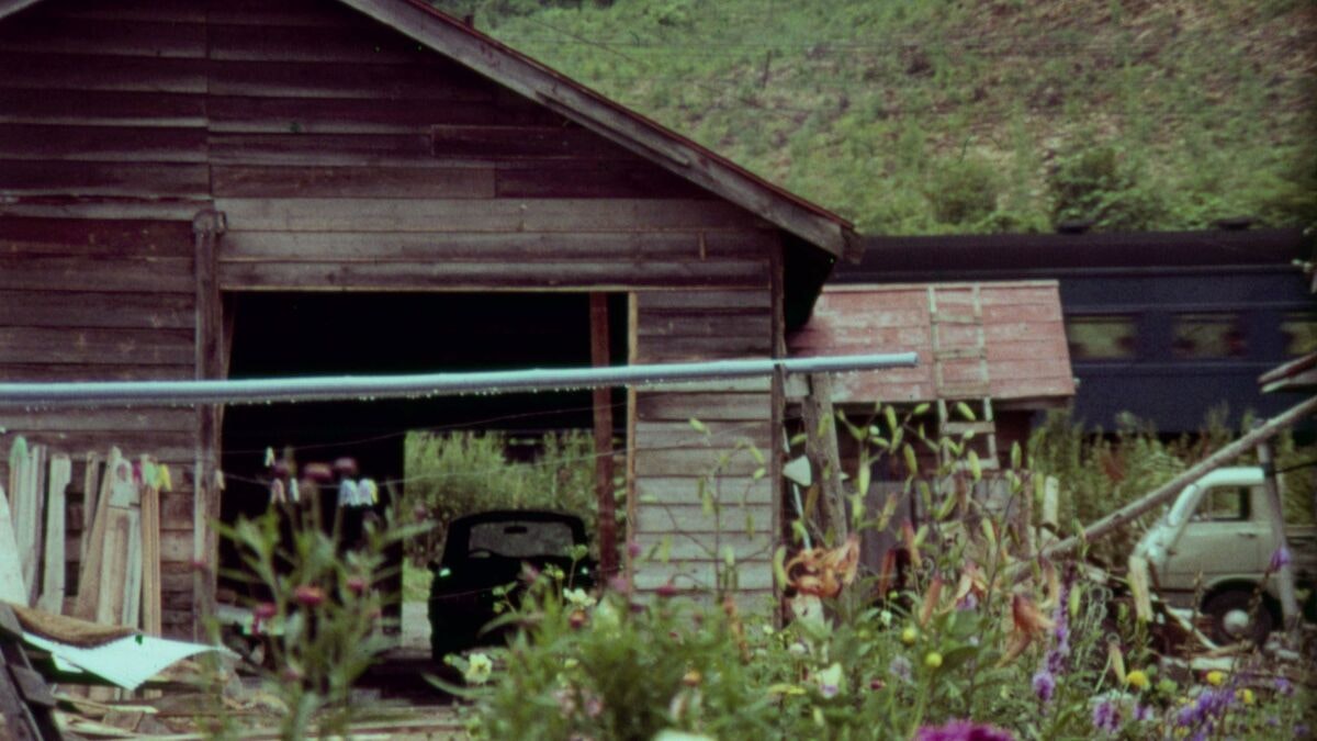 A film still of a wooden shack with a truck, flowering garden and washing line out front, while a vehicle sits in the garage. Slats of timber surround the shack.