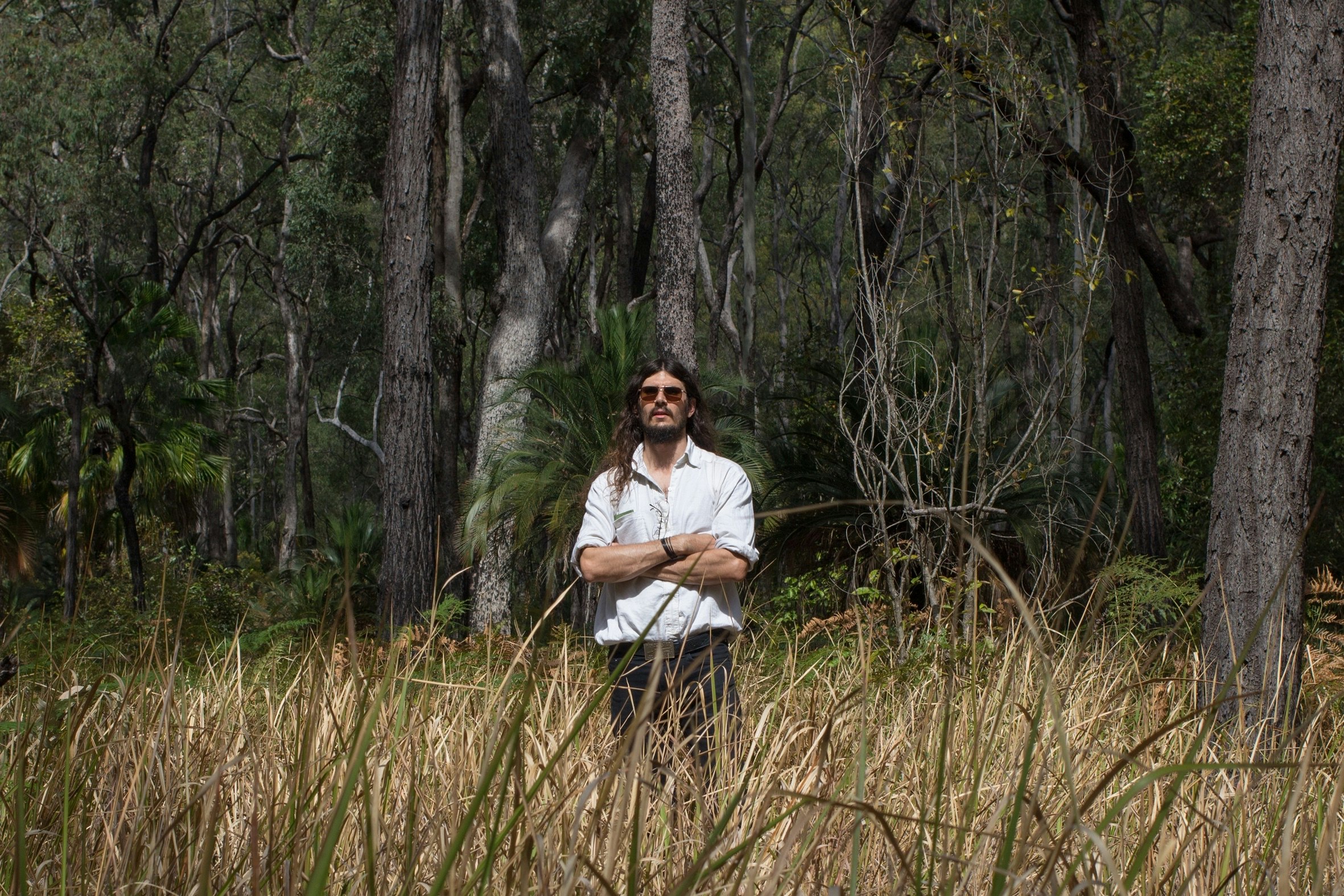 A male-presenting figure with long brunette hair and sunglasses stands with his arms folded in a paddock with grass rising up to his hips.