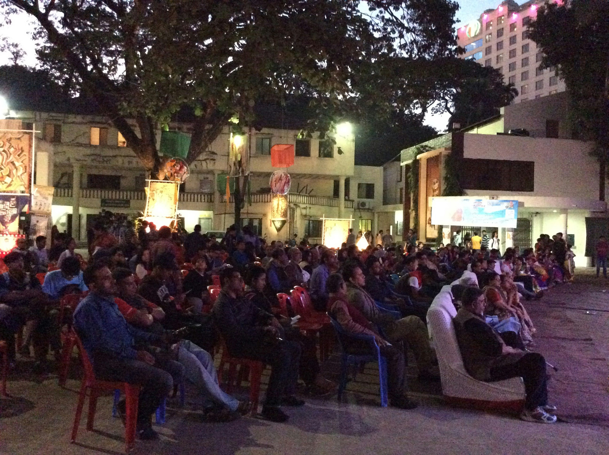 A Bangladeshi audience sits on chairs in an outdoor setting in the evening, looking at an illuminated production (production is out of frame).