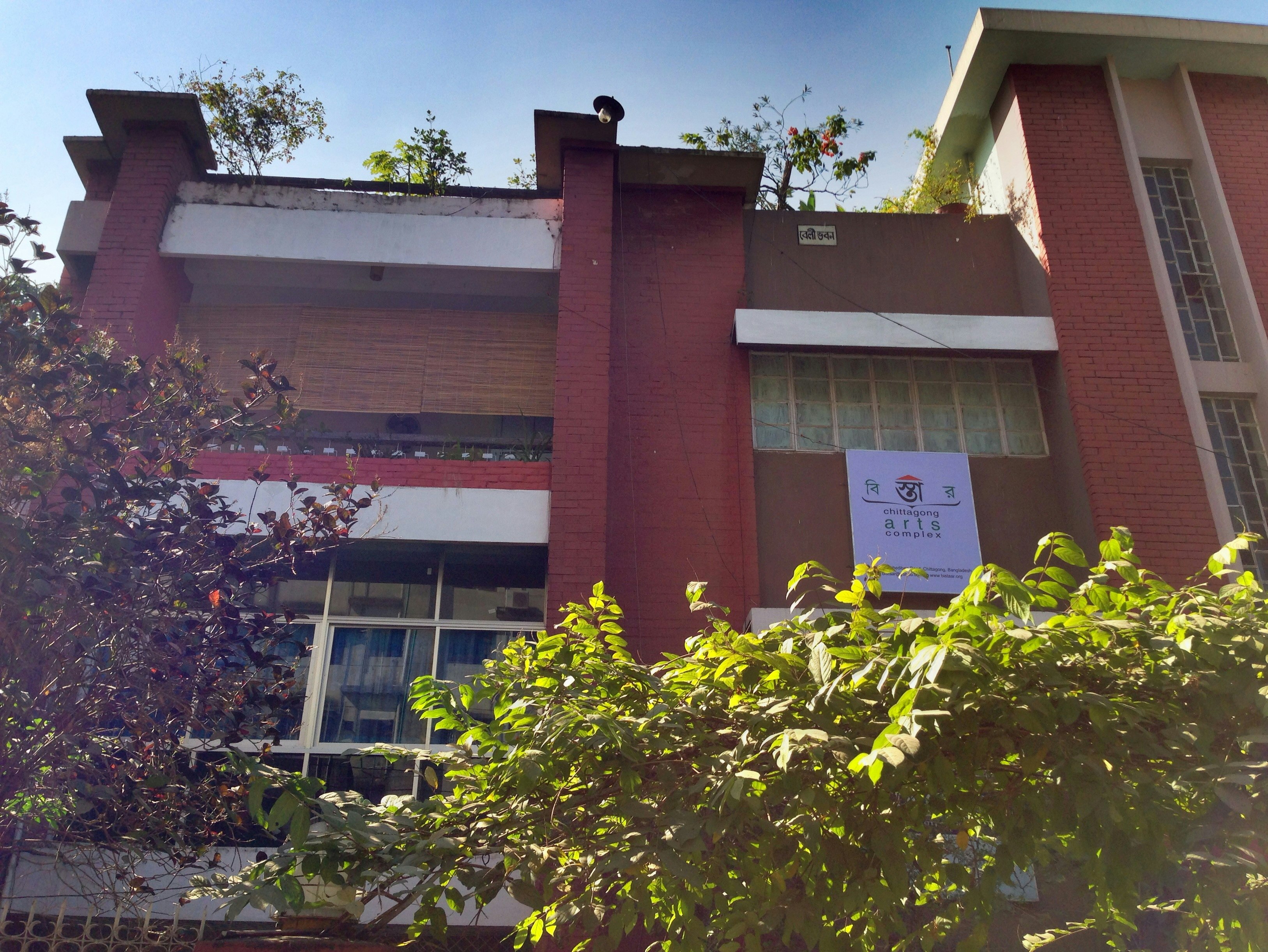 A facade of a red brick and concrete building, with the sign 'Chittagong Arts Complex' installed under one window.