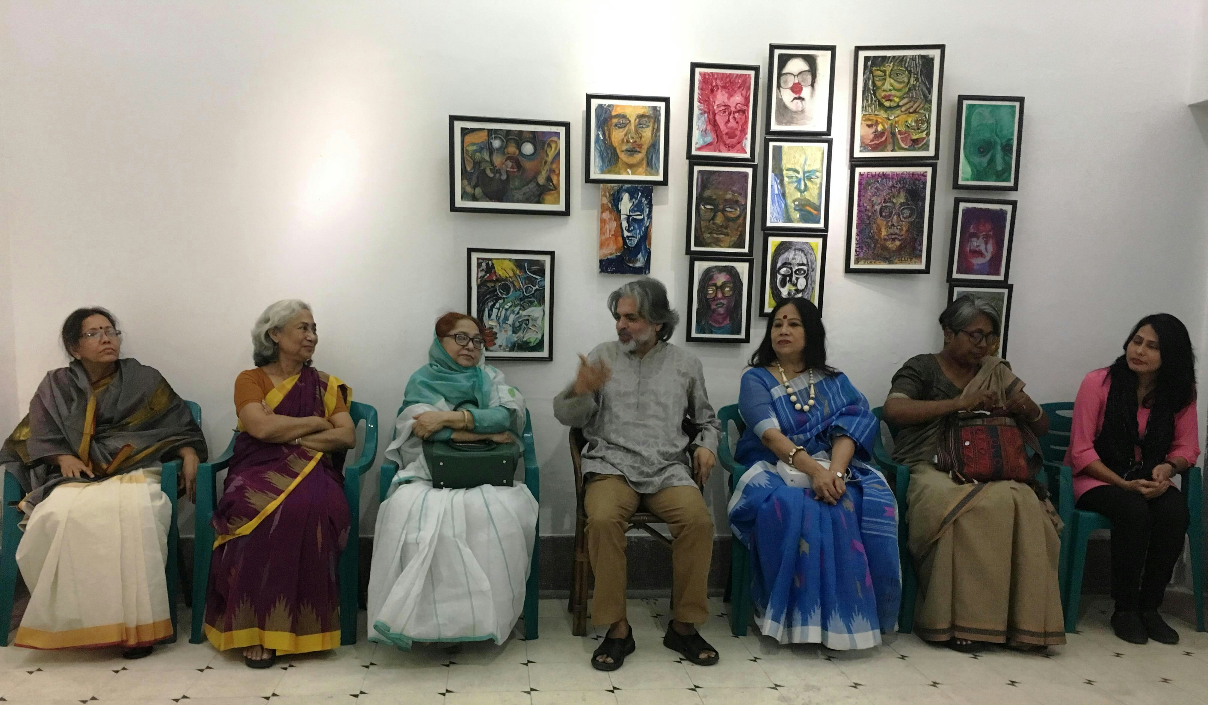 Six female-presenting brown women in saris seated against a wall, with Alam Khorshed seated in the middle talking to them. A series of colourful illustrations are hanging behind them on the wall.