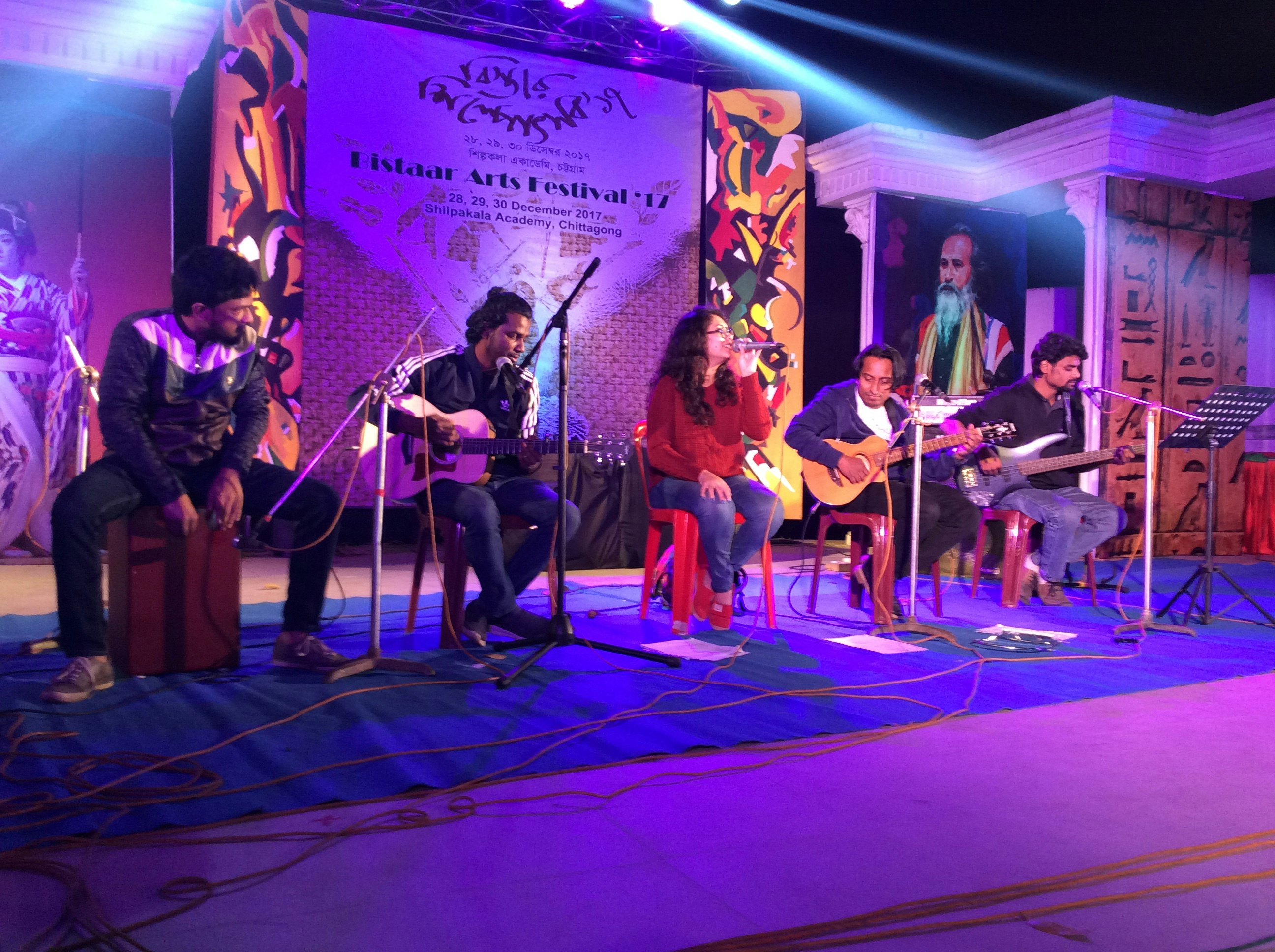 Five seated musicians performing on a stage lit with purple light. One of them is drumming on his stool, three are playing guitars and one is singing into a microphone.