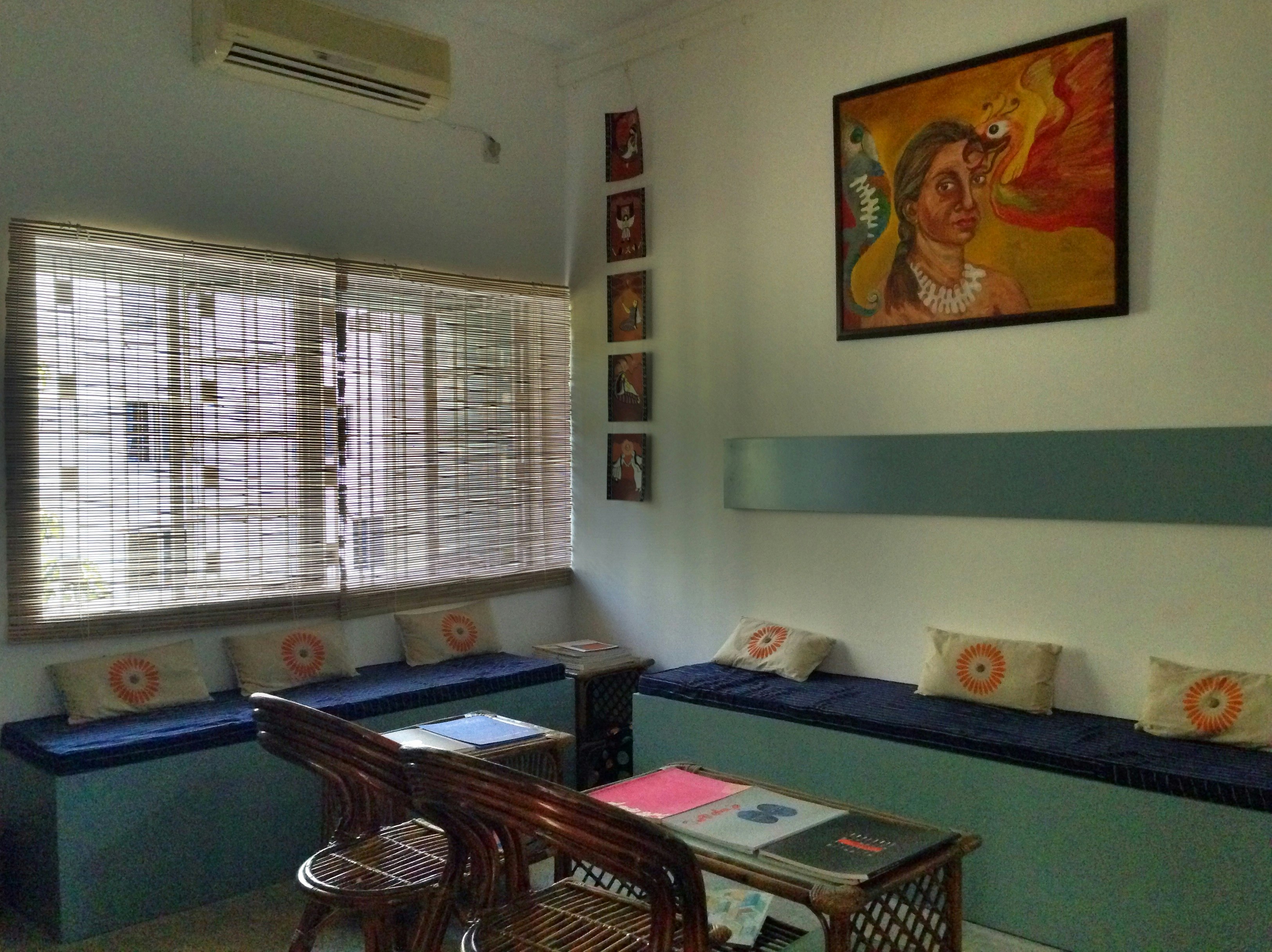 Two cushioned benches in a corner of the arts complex, facing some wooden chairs and coffee tables with art books spread on them. On the wall is a painting of a brown-skinned female-presenting figure staring out with a red flamingo next to her.