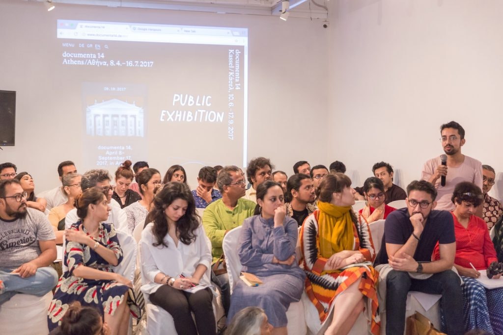 A male-presenting figure holding a microphone stands up amidst a seated art gallery audience. On the wall behind him is a projection that reads, 'documenta 14: Athens/PUBLIC EXHIBITION'