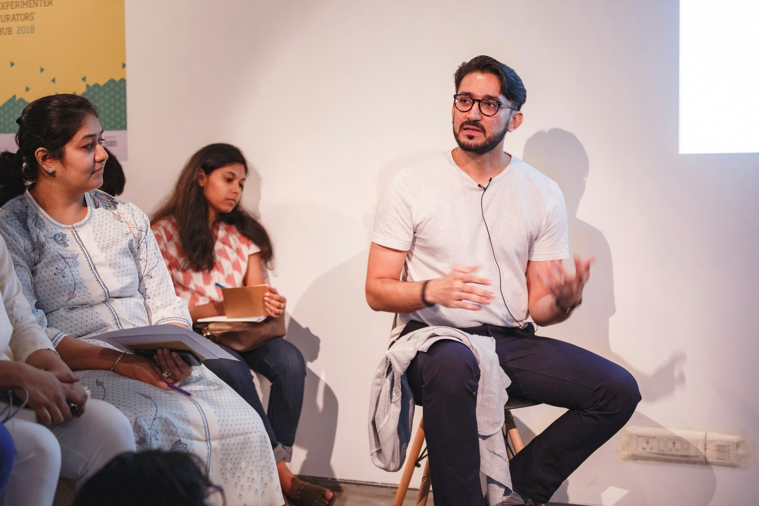 A male-presenting figure wearing glasses, a white t-shirt and black trousers sits on a stool, talking and gesturing to a crowd out of frame.