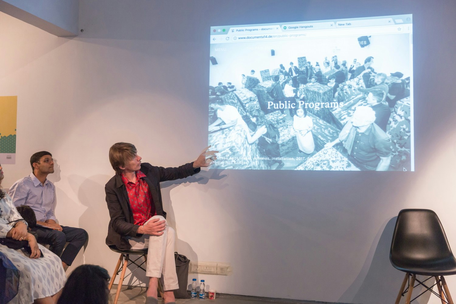 A male-presenting figure wearing a printed red button-up shirt and a black blazer and white trousers gestures to a projection of the documenta 14 website on the wall.