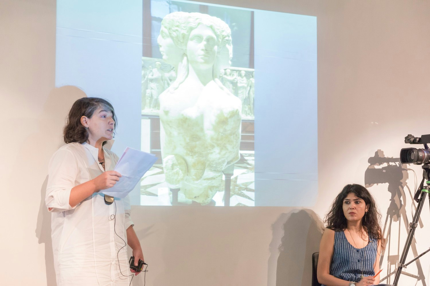A female-presenting figure in a long white button-up shirt holding some sheets of white paper speaks in front of a projection of a three-headed bust.