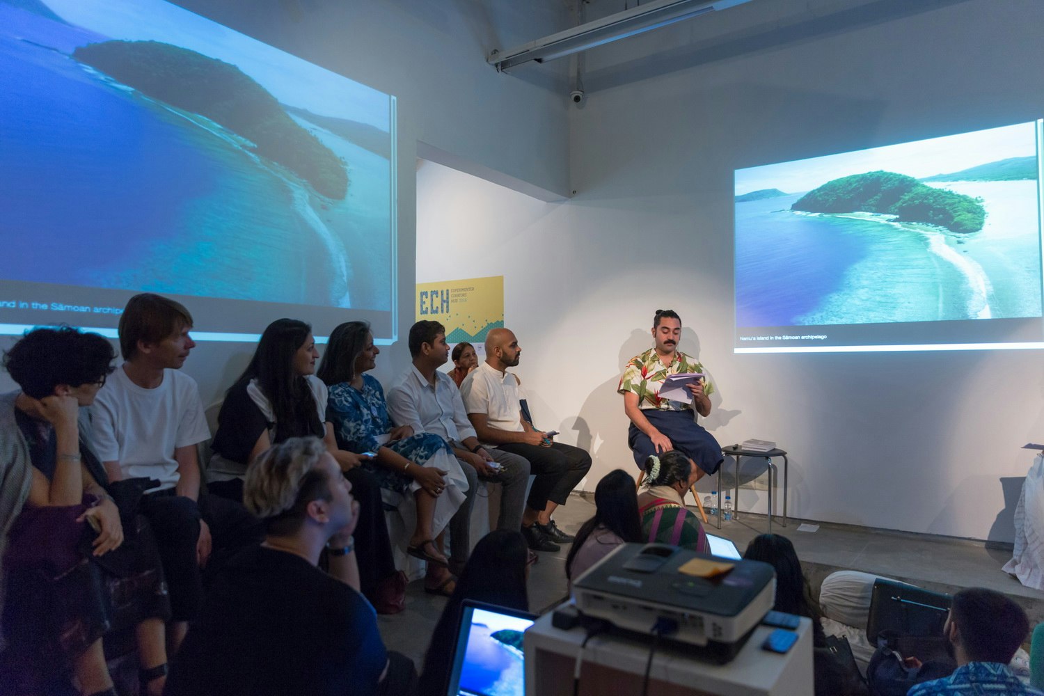 A Pasifika male-presenting figure with a black moustache and hair pulled back into a bun reads from a sheet of paper, while seated in front of a projection of an island in the Samoan archipelago. An audience sitting in the shadows looks on.