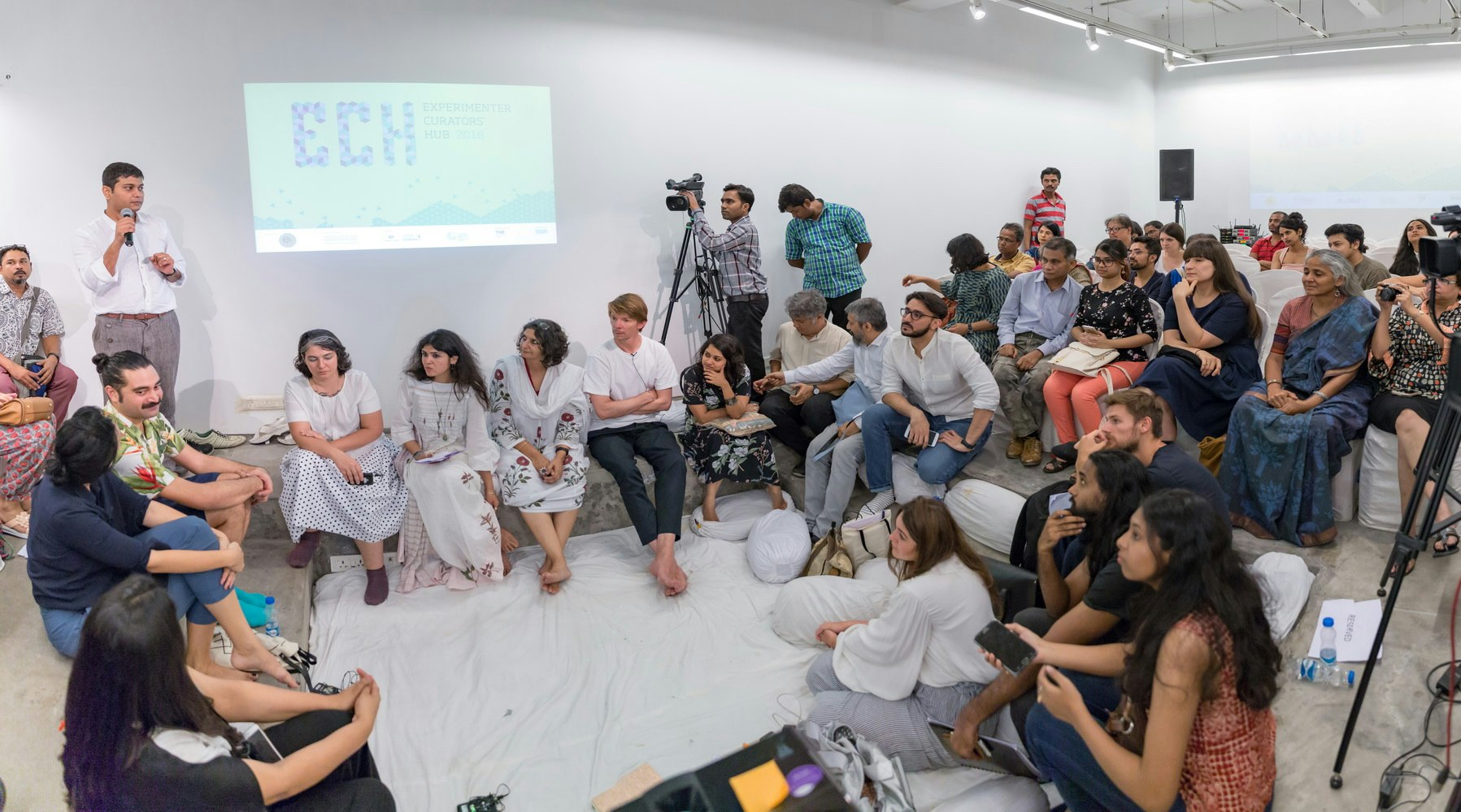 A seated crowd in a white art gallery space laid with a drop sheet, looking upon a brown-skinned male-presenting figure in a white dress shirt and grey trousers as he speaks into a microphone.