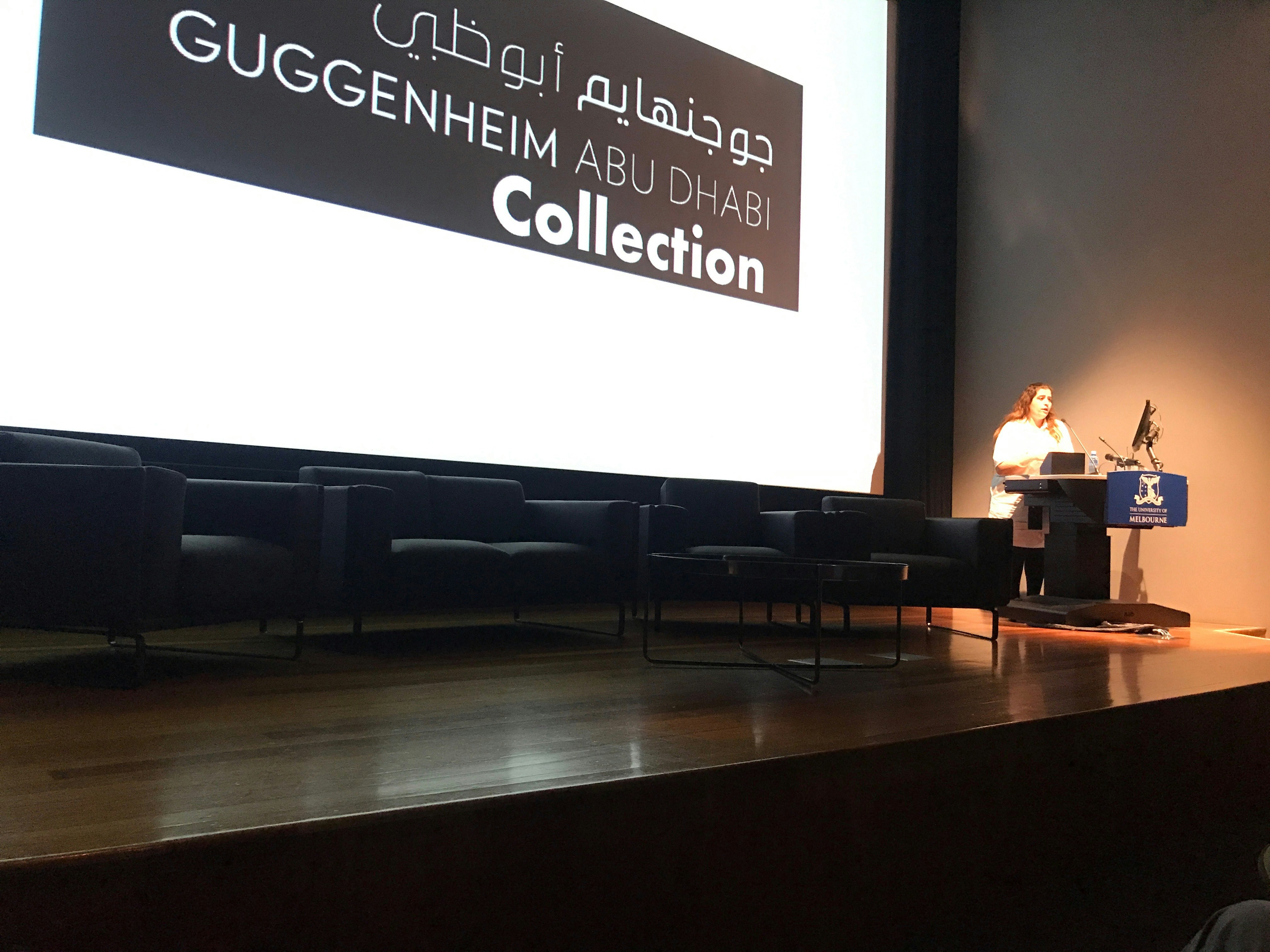 A Middle Eastern female-presenting figure stands at a podium decorated with The University of Melbourne insignia. Behind her is a large projector screen that reads: Guggenheim Abu Dhabi Collection.