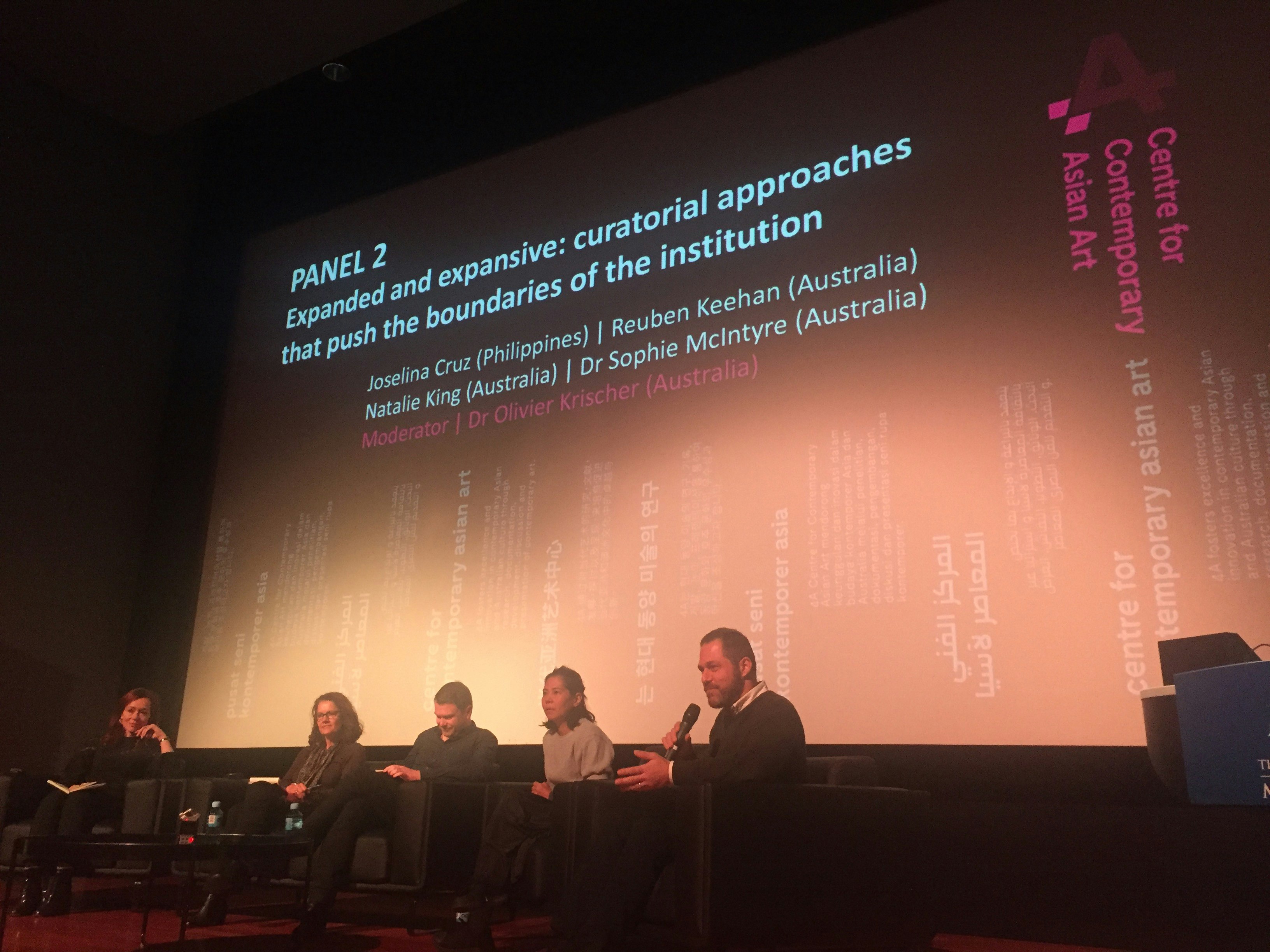 Four panellists sit in sofas next to a moderator, in front of a large projector screen that reads: Panel 2: Expanded and expansive: curatorial approaches that push the boundaries of the institution.