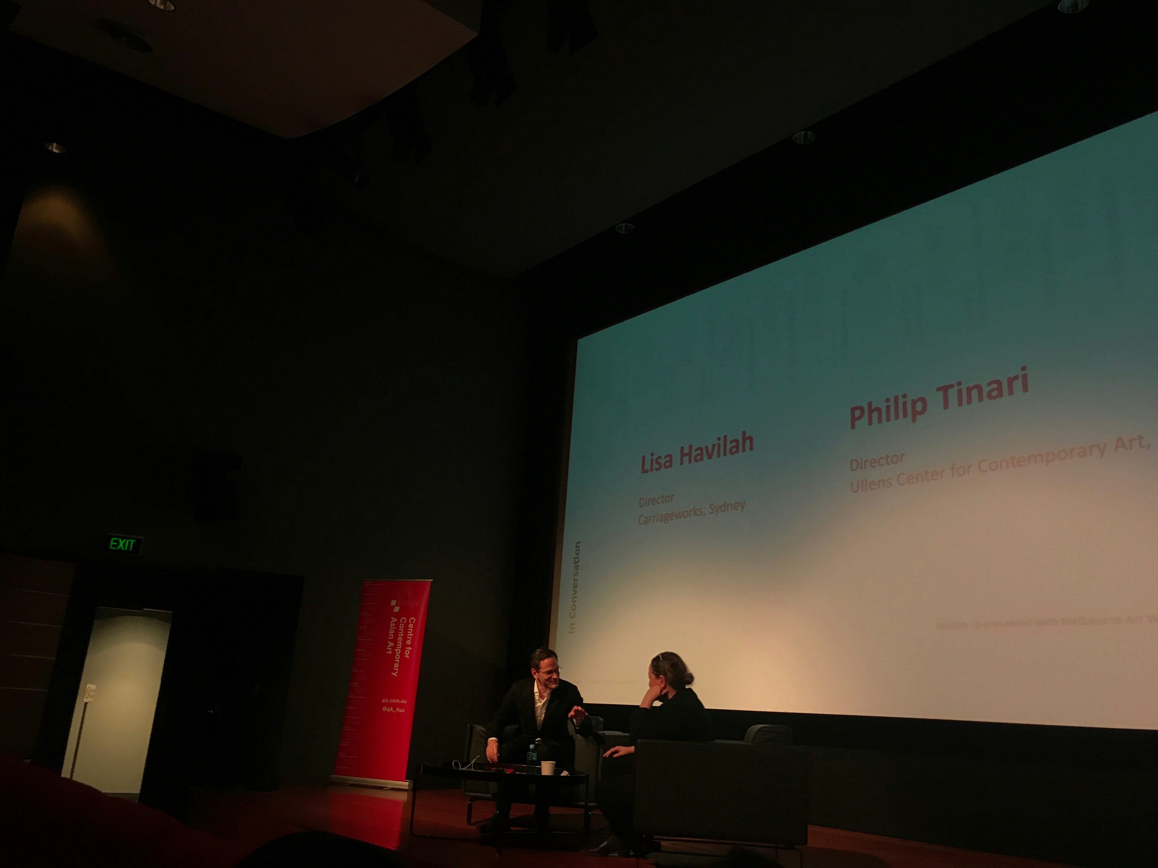 Two figures sitting on stage face each other in conversation, while the large projector screen behind them reads: Lisa Havilah, Philip Tinari.