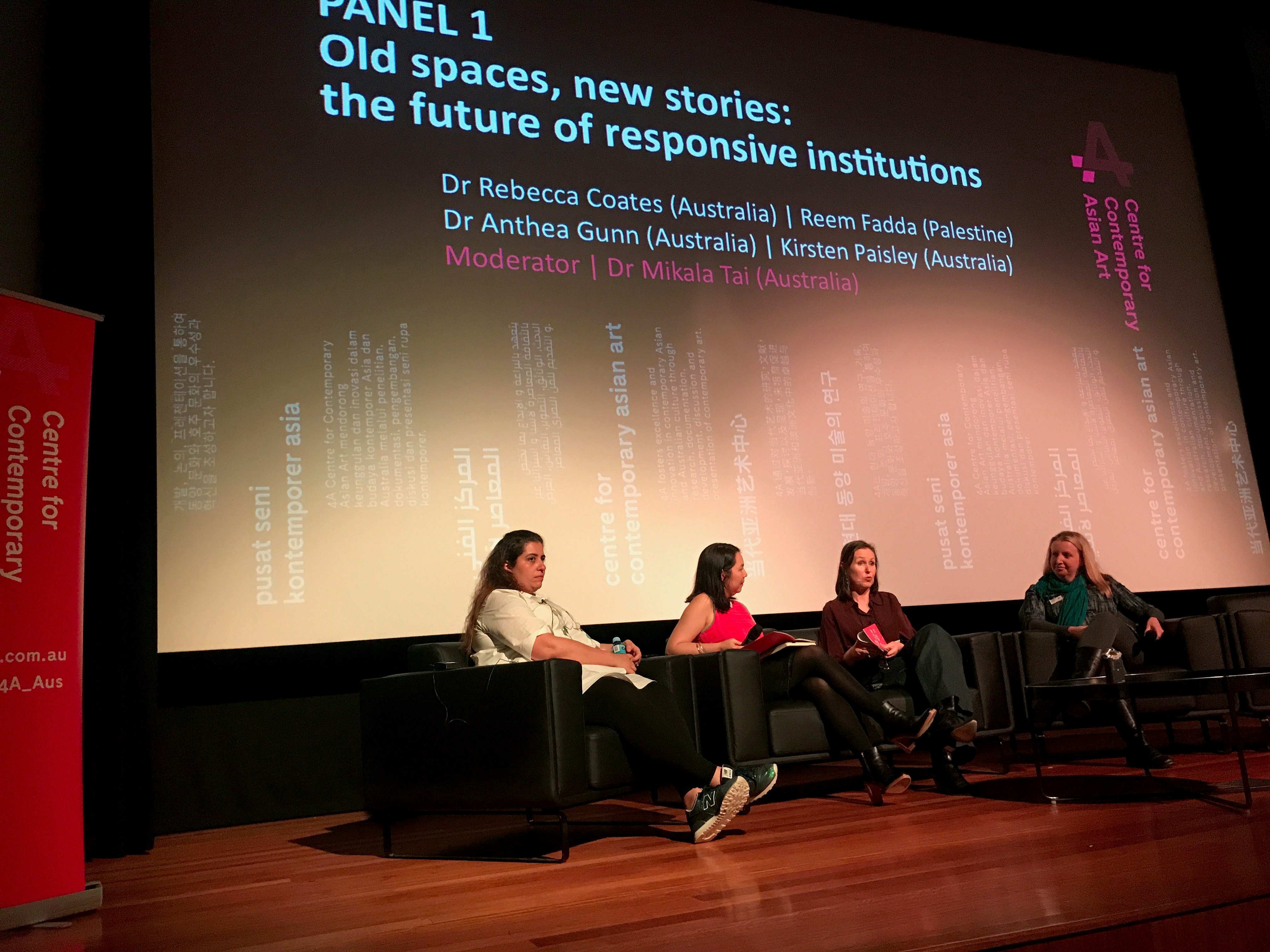 Four female-presenting figures seated on a stage, behind them a large projector screen that says, Panel 1: Old spaces, new stories: the future of responsive institutions.