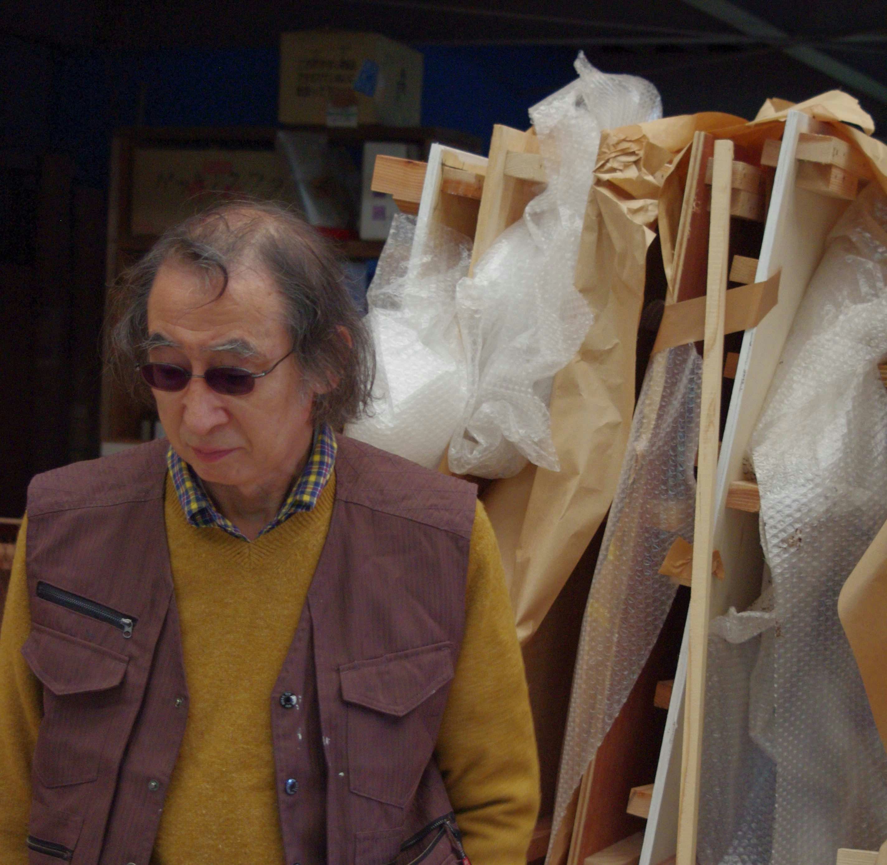 An elderly Japanese male-presenting figure with grey hair, dark glasses and wearing a checked shirt, yellow sweater and maroon vest stands in his studio, looking downwards at the floor.