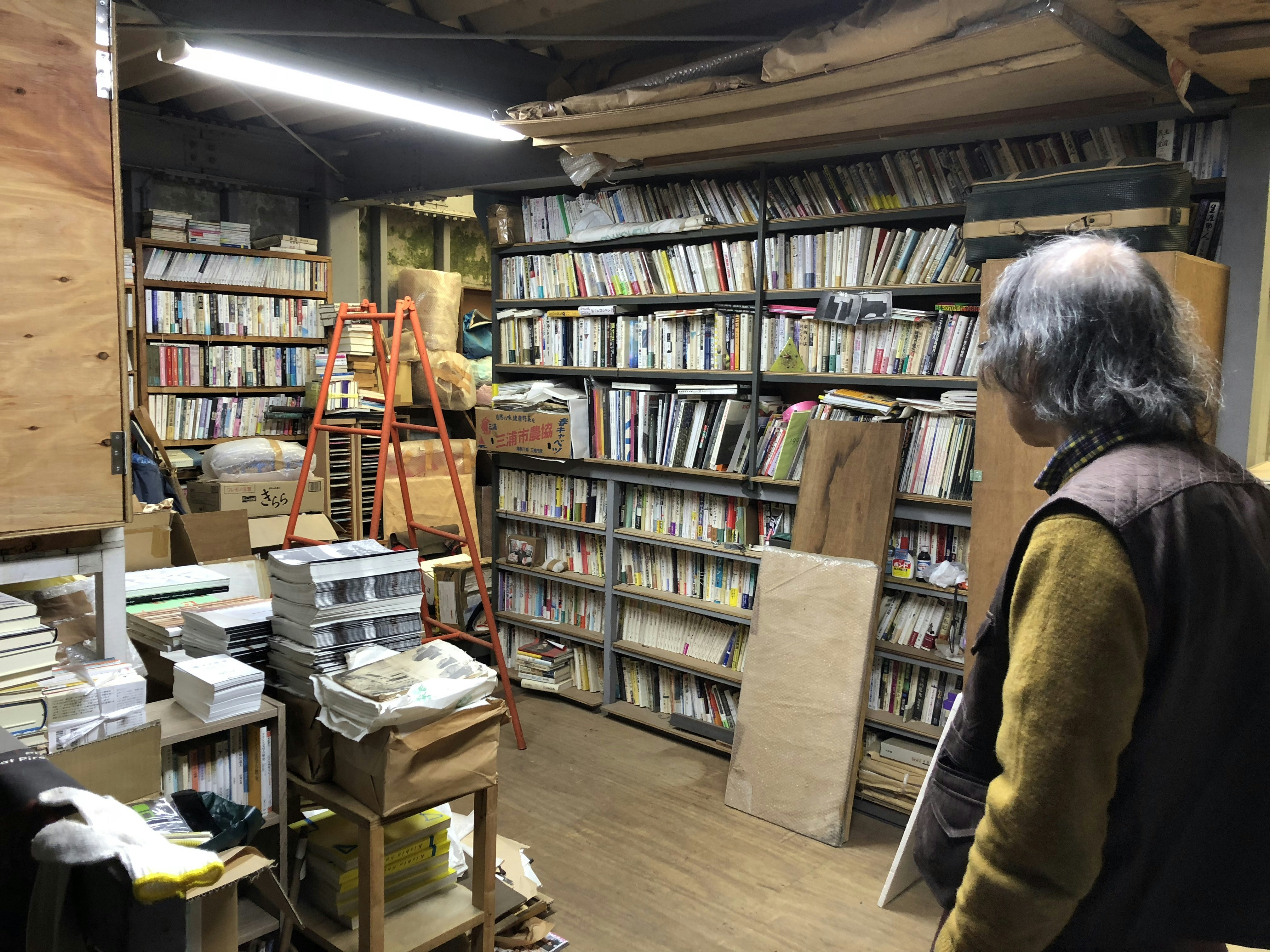 Kishio Suga stands with his back to us, looking at a library of stacked books, magazines and newspapers in boxes, next to planks of wood, wrapped packages and an orange stepladder.